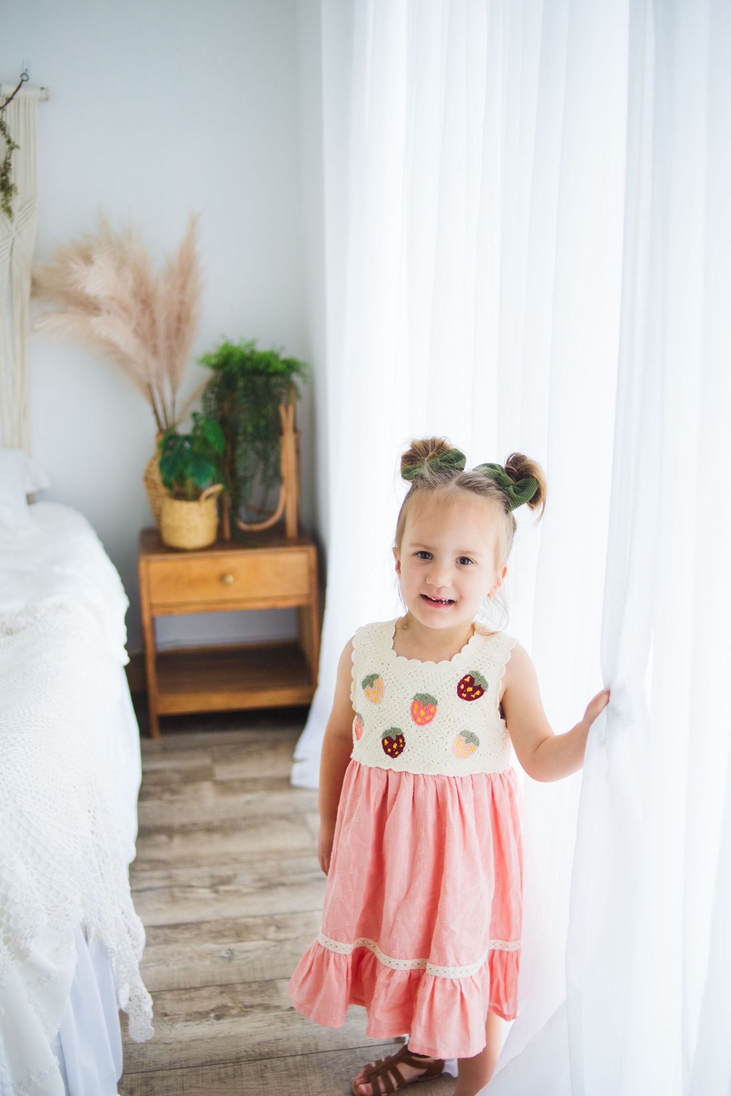 A toddler wearing a pink and white dress with fruit patterns stands in a bright bedroom.