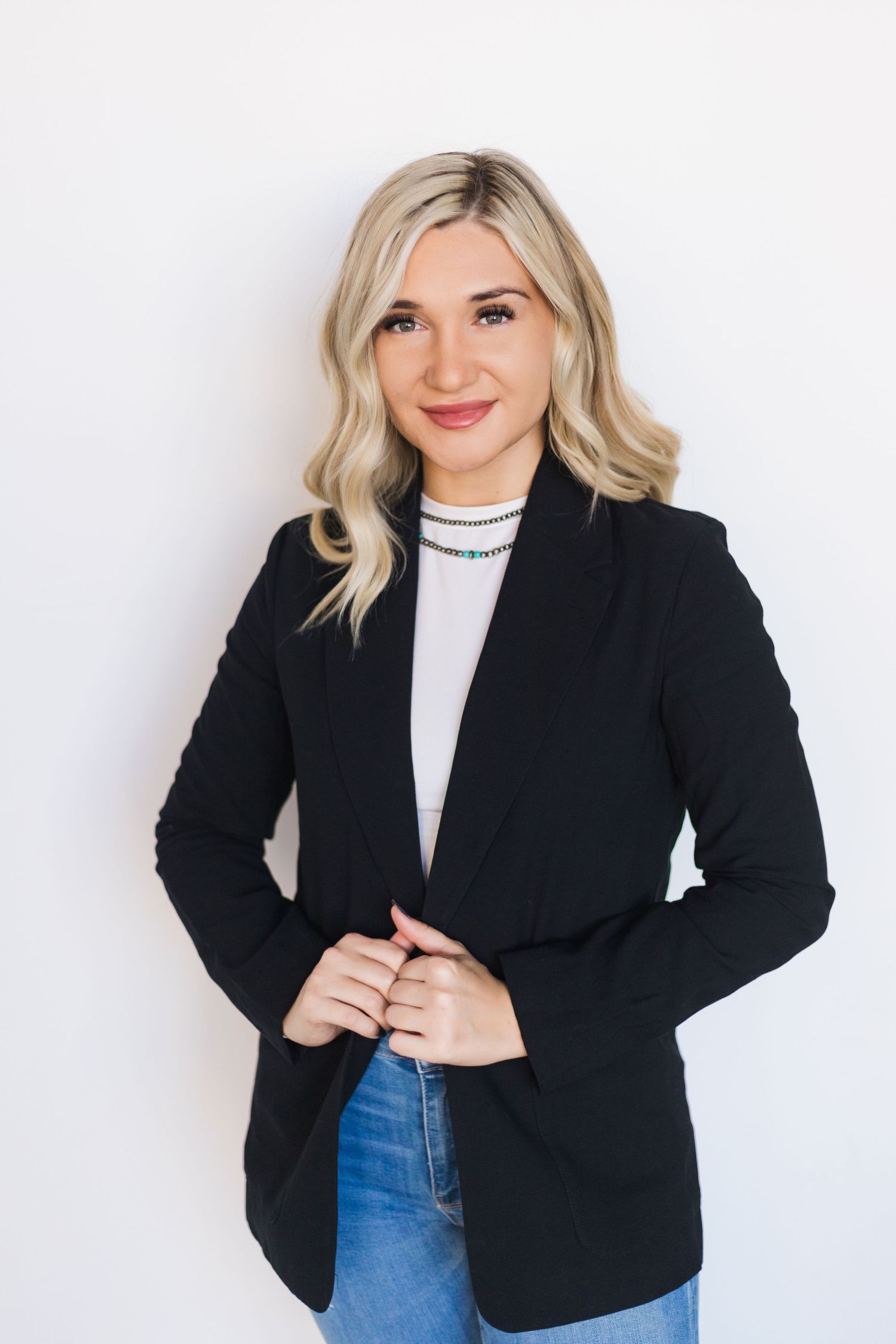 Woman models black blazer with white top and jeans in studio setting.