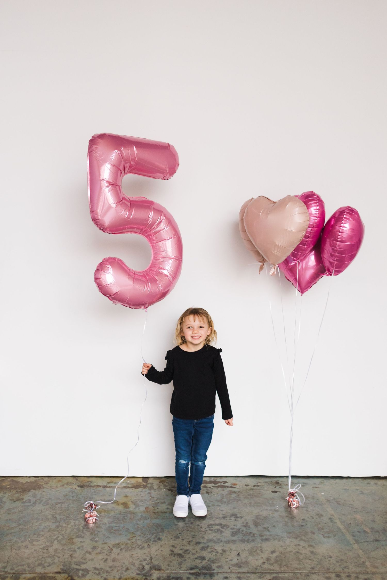 Pink number 5 balloons float beside child in casual outfit against white background.