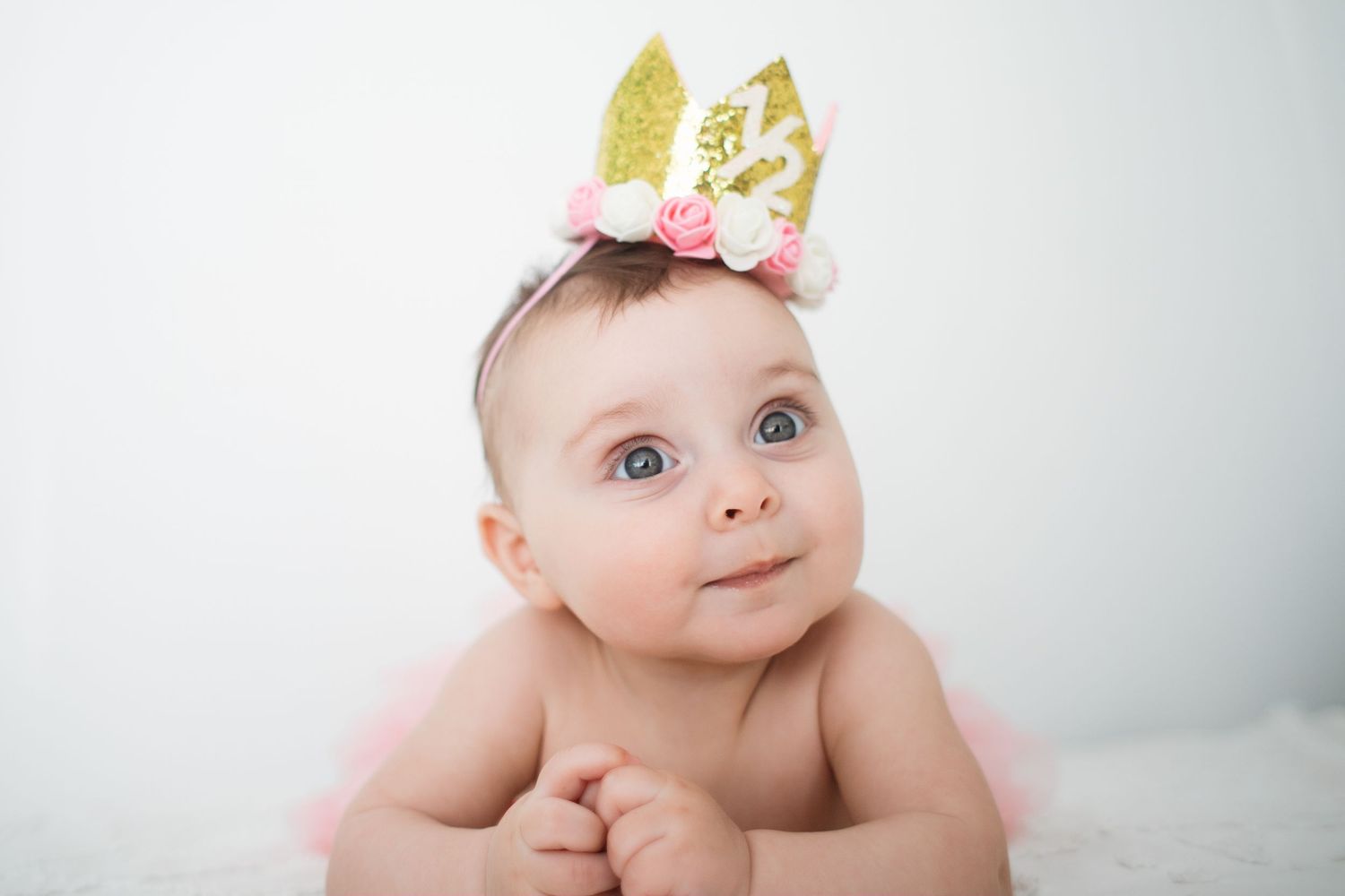 Adorable baby wearing a gold glitter crown with pink roses, looking up with blue eyes on a white background.