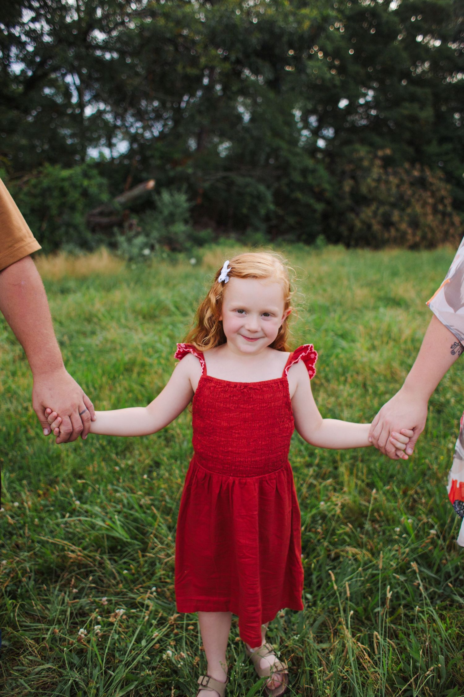 Young child in red dress holding hands with adults while walking outdoors.