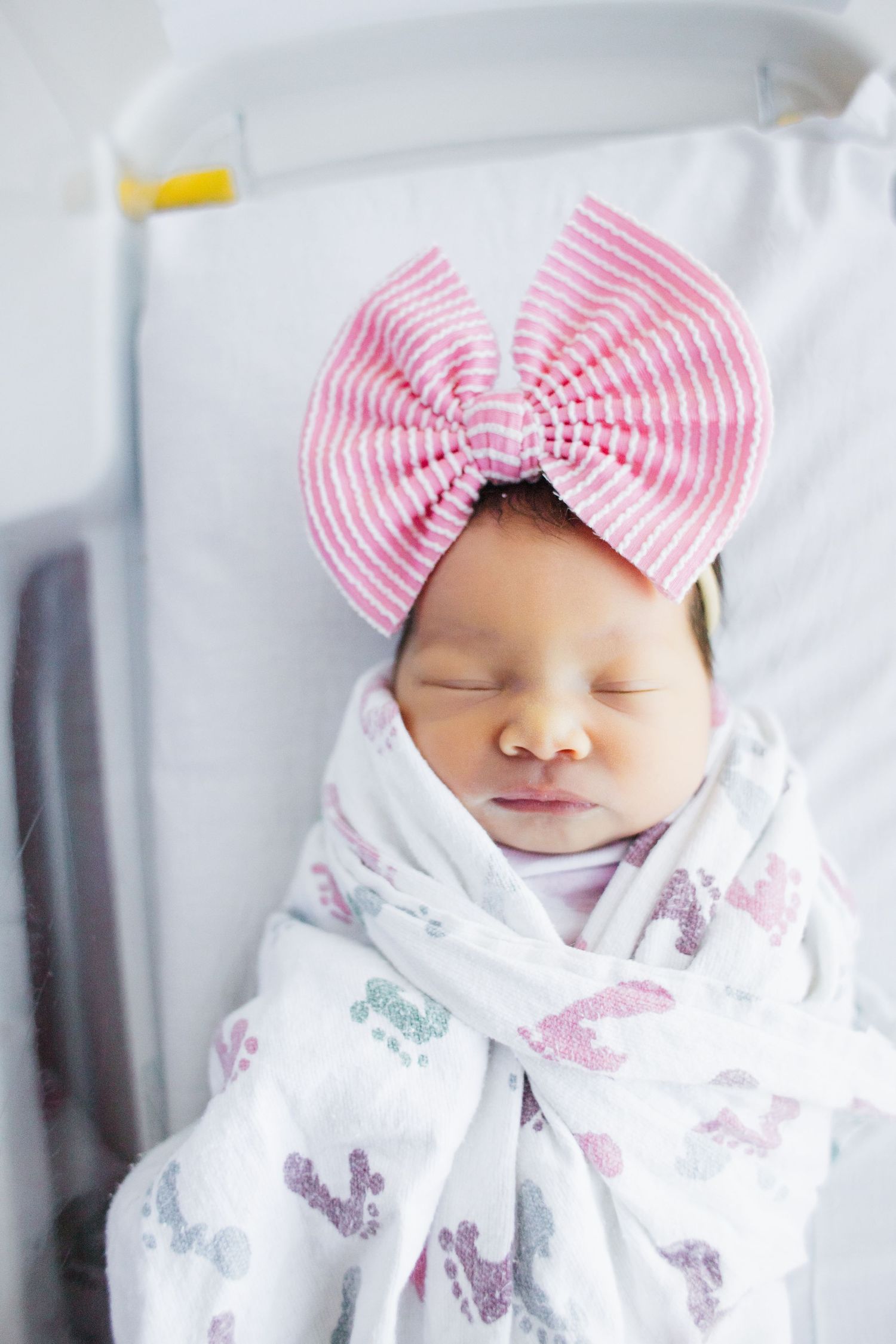 Newborn baby wearing white swaddle with butterfly print and pink striped bow while sleeping in peaceful repose.