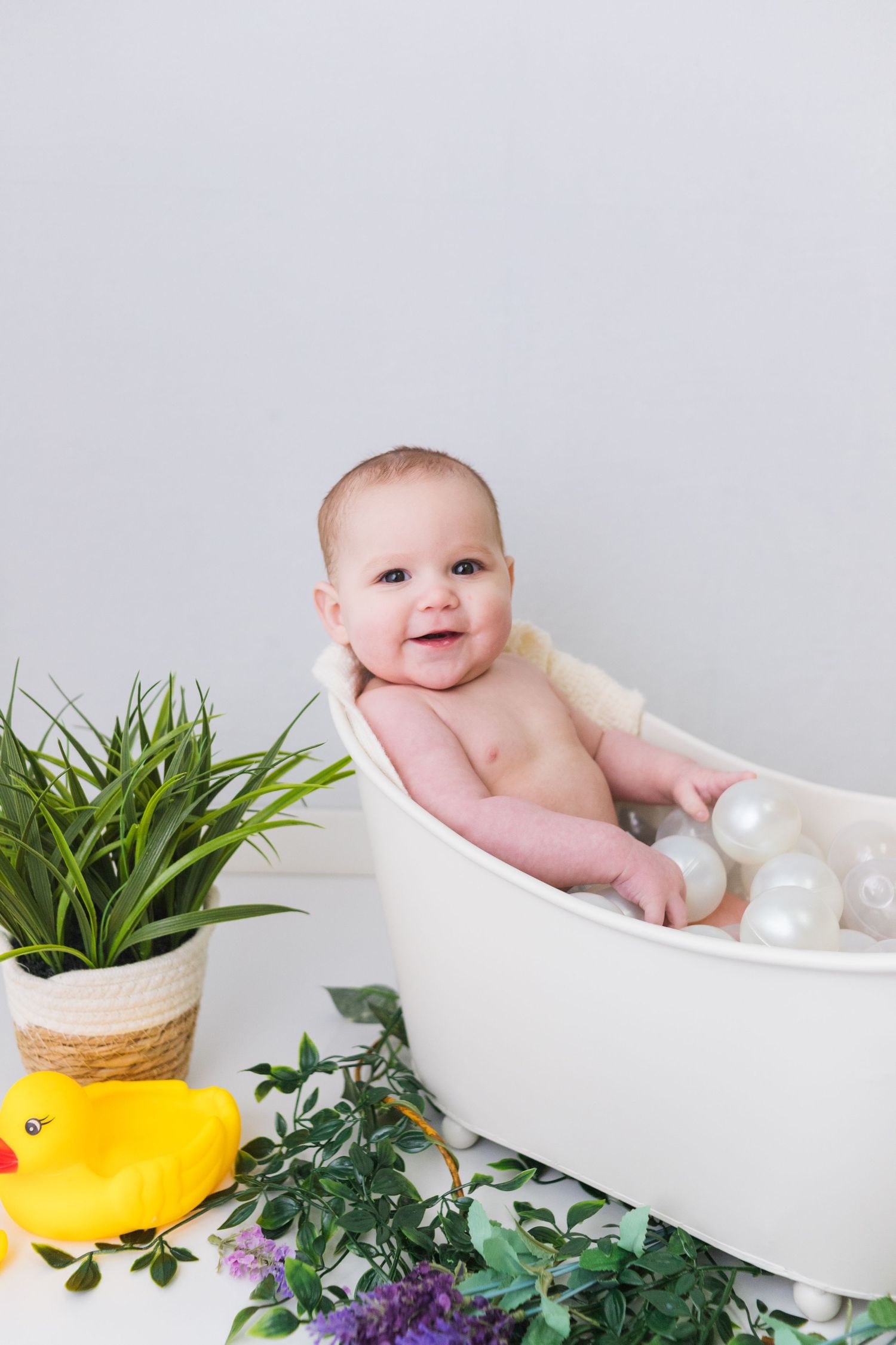 Baby bath photography setup with white tub, potted plants, and purple flowers.