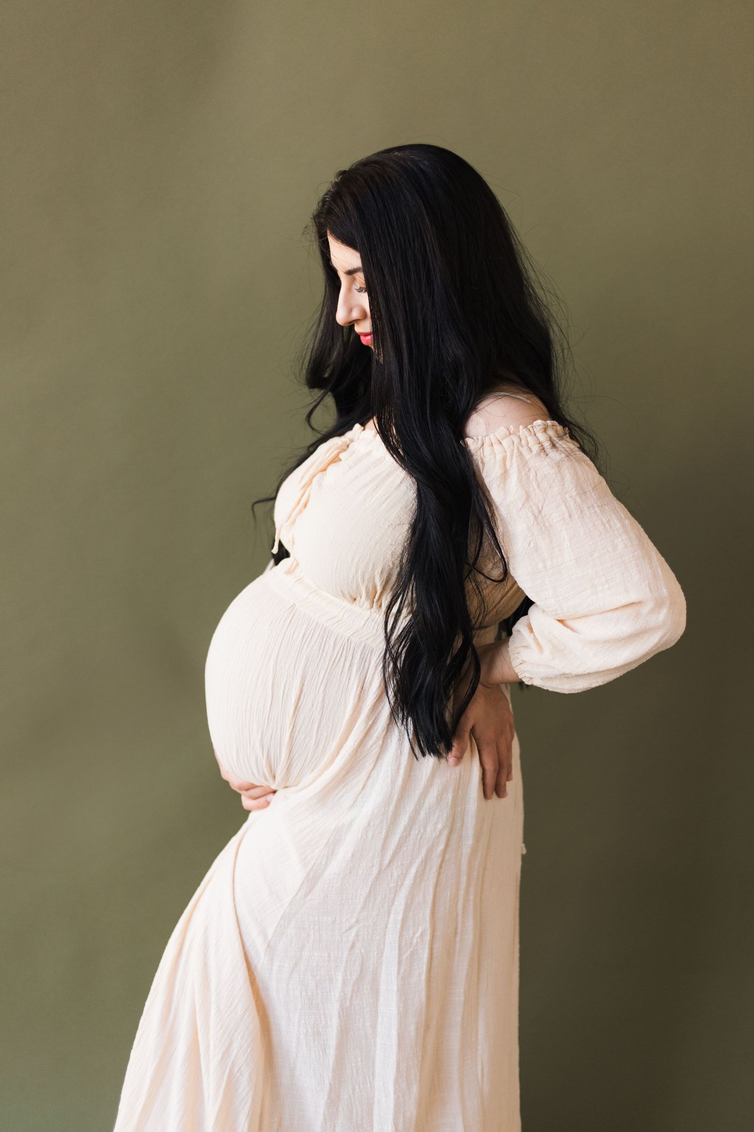 A maternity photoshoot featuring flowing cream dress against sage green wall.