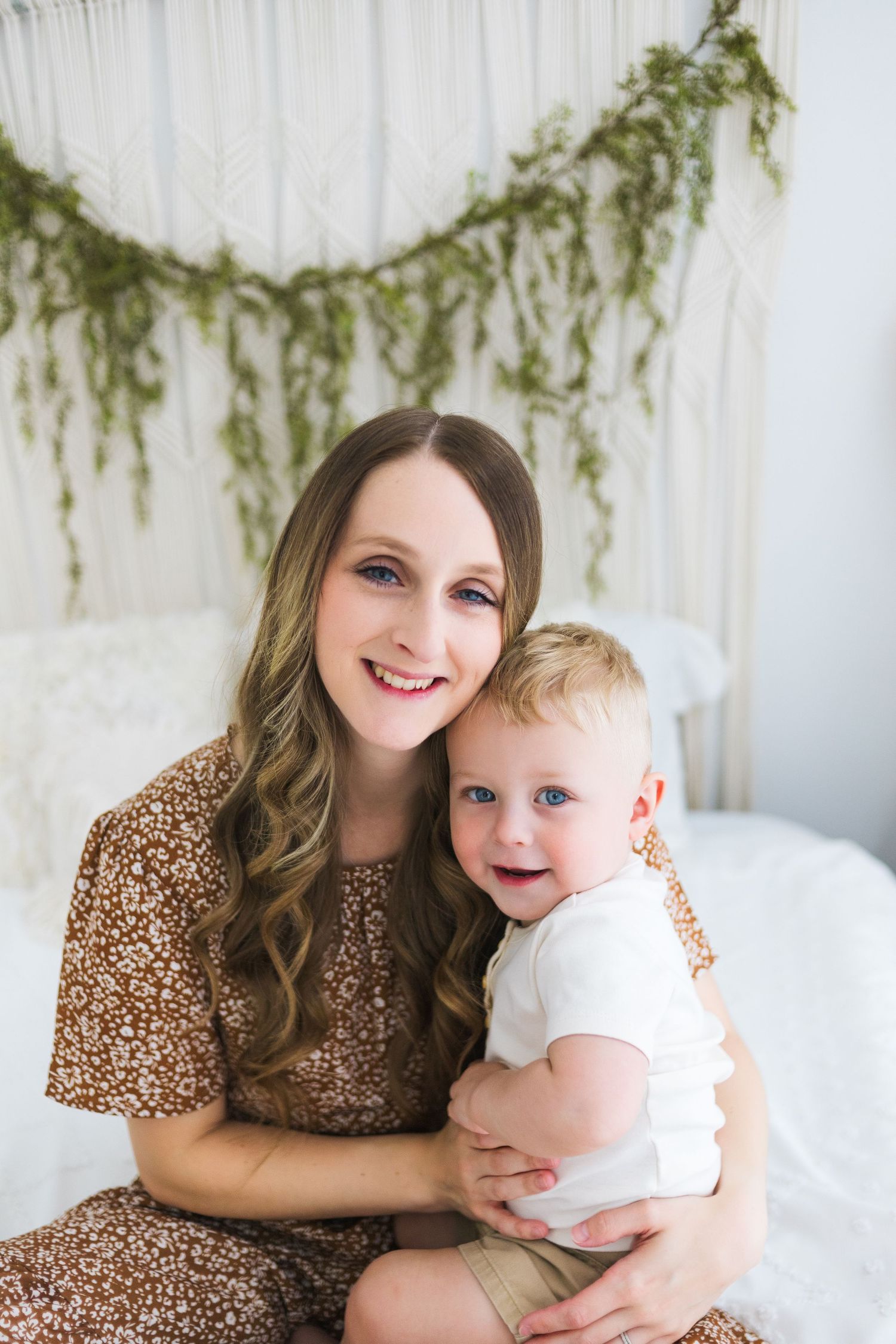 A tender moment captured between mother and child sitting together against a white backdrop with greenery decor.