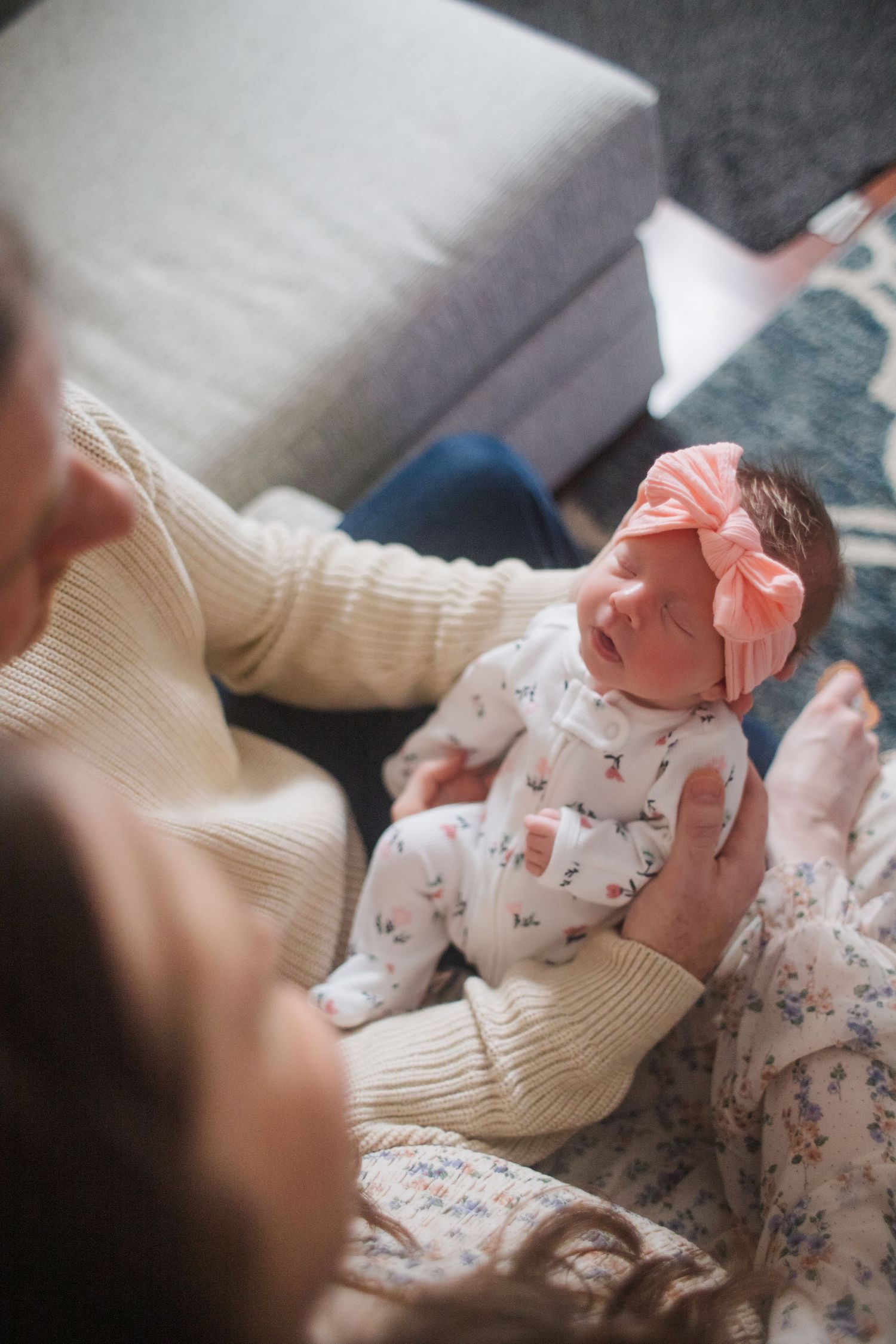 A sleeping baby wearing a white floral onesie and coral bow headband rests peacefully on cream-colored bedding.