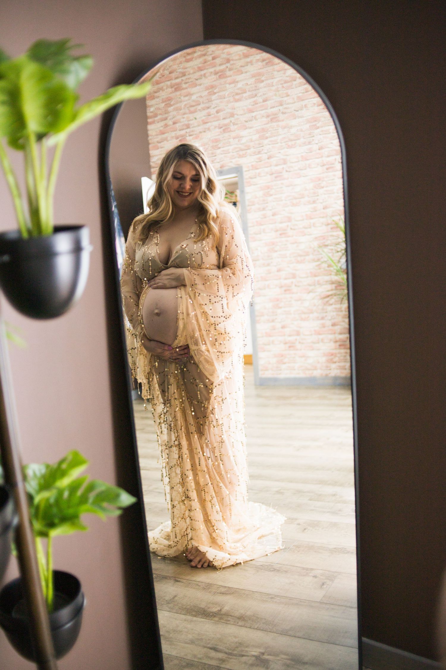 Mirror reflection shows expectant figure in elegant lace gown with indoor plants.