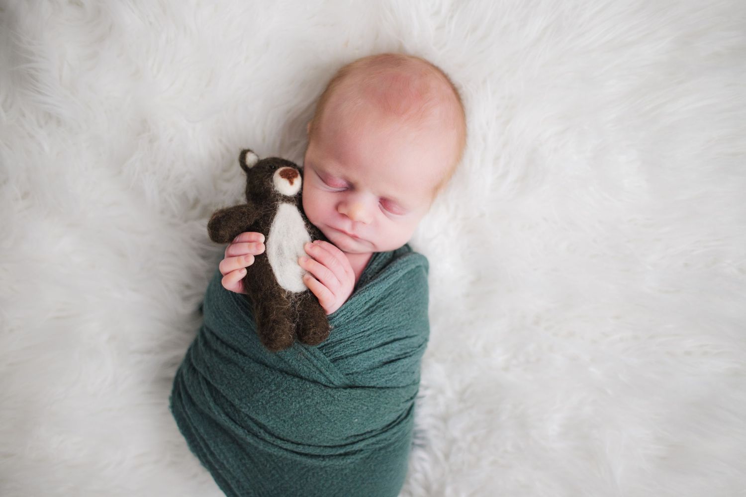 A newborn baby swaddled in green fabric lies peacefully on a soft white blanket during a professional photo session.
