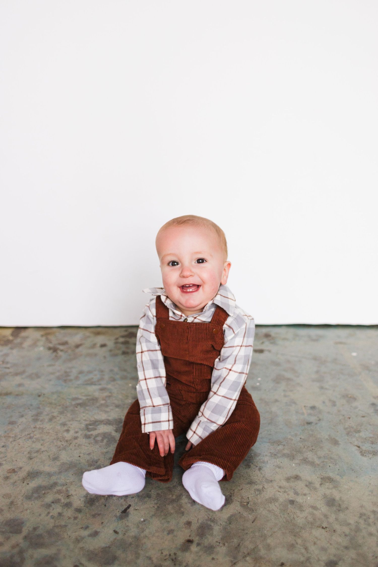 Baby in brown overalls and plaid shirt sits happily on concrete floor.
