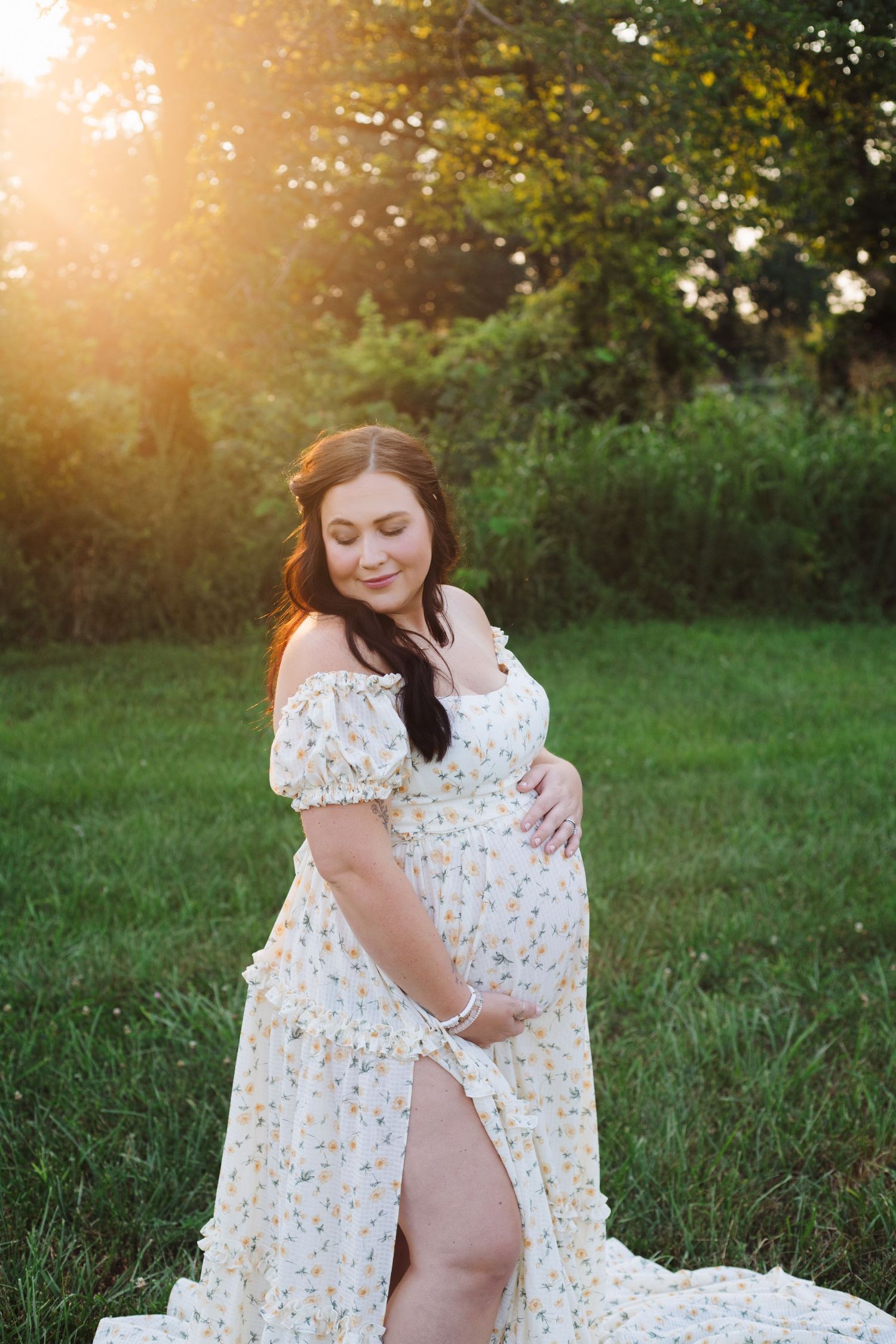 A woman in a floral dress stands in a sunlit meadow during golden hour with soft glowing light filtering through trees.