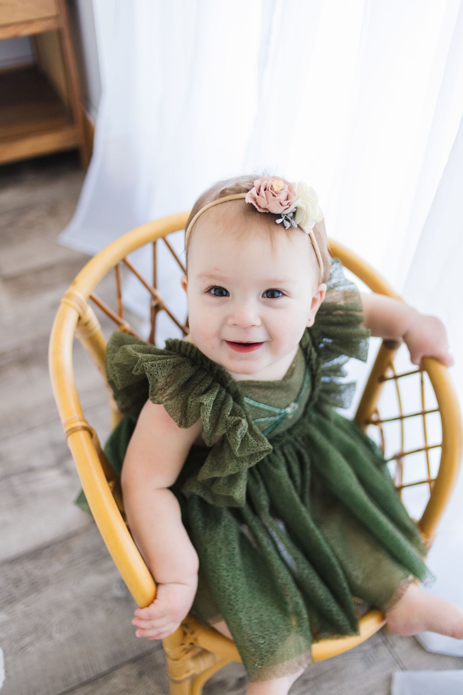 A baby wearing a green ruffled dress and floral headband sits smiling in a rattan chair against white curtains.