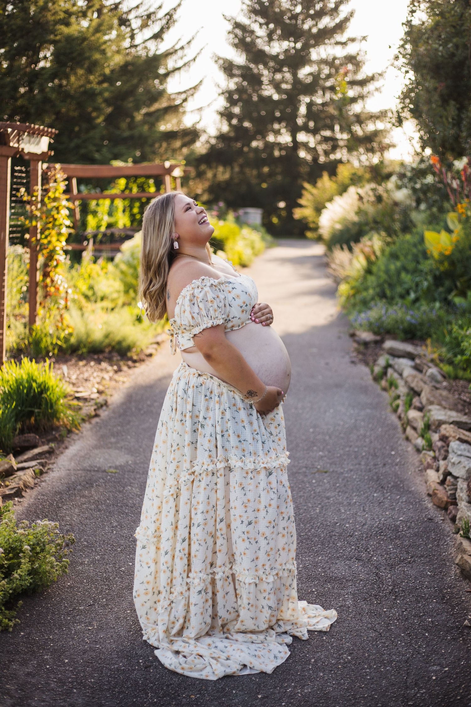 Expectant woman in flowing white lace dress stands on garden path during golden hour sunset.