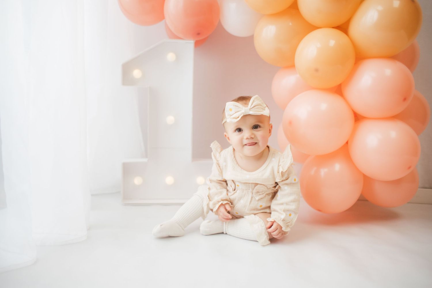 A baby in a white dress sits on a floor with peach-colored balloons and white decor in the background.