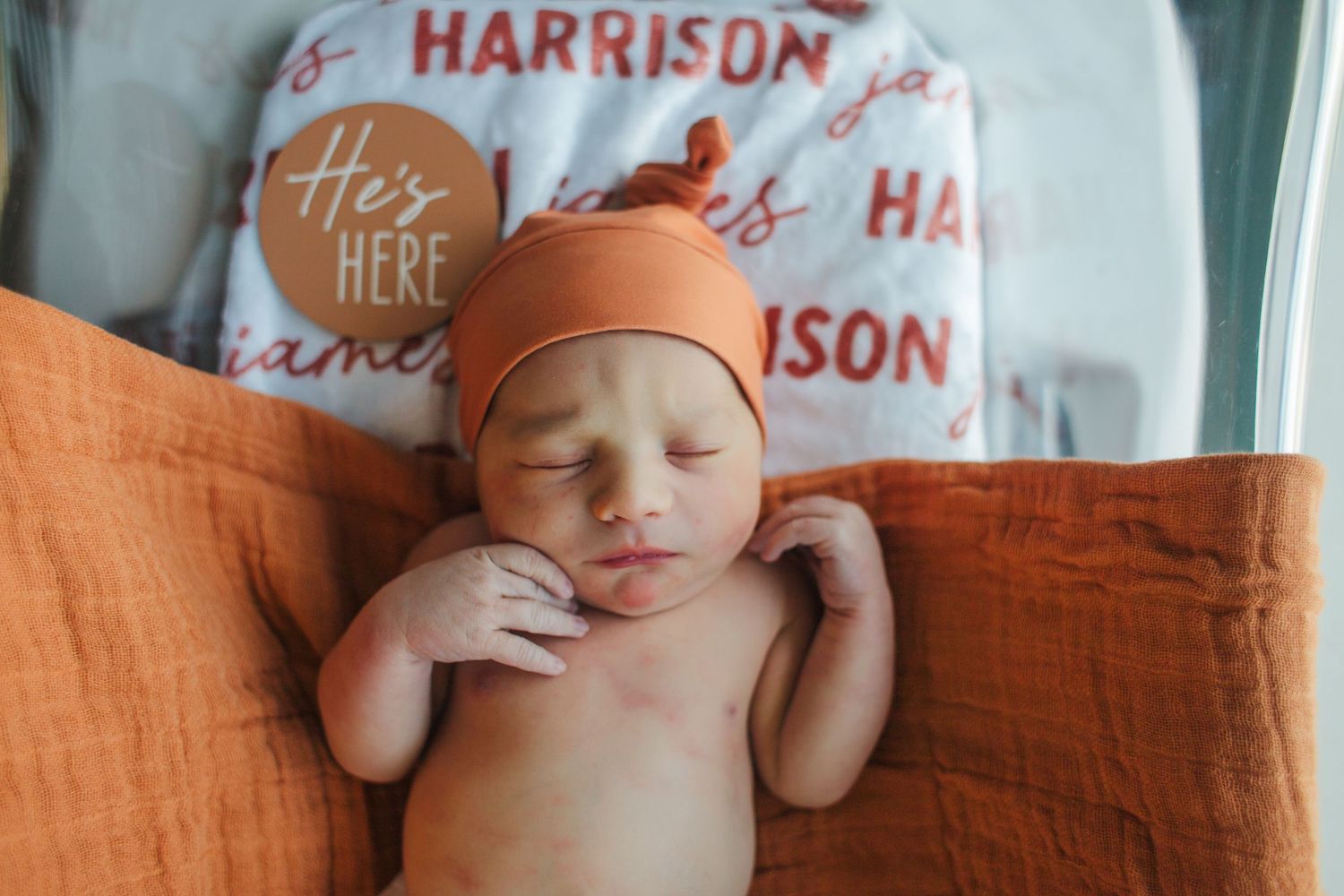 A sleeping newborn baby wearing an orange knit hat lies on a peach-colored blanket.