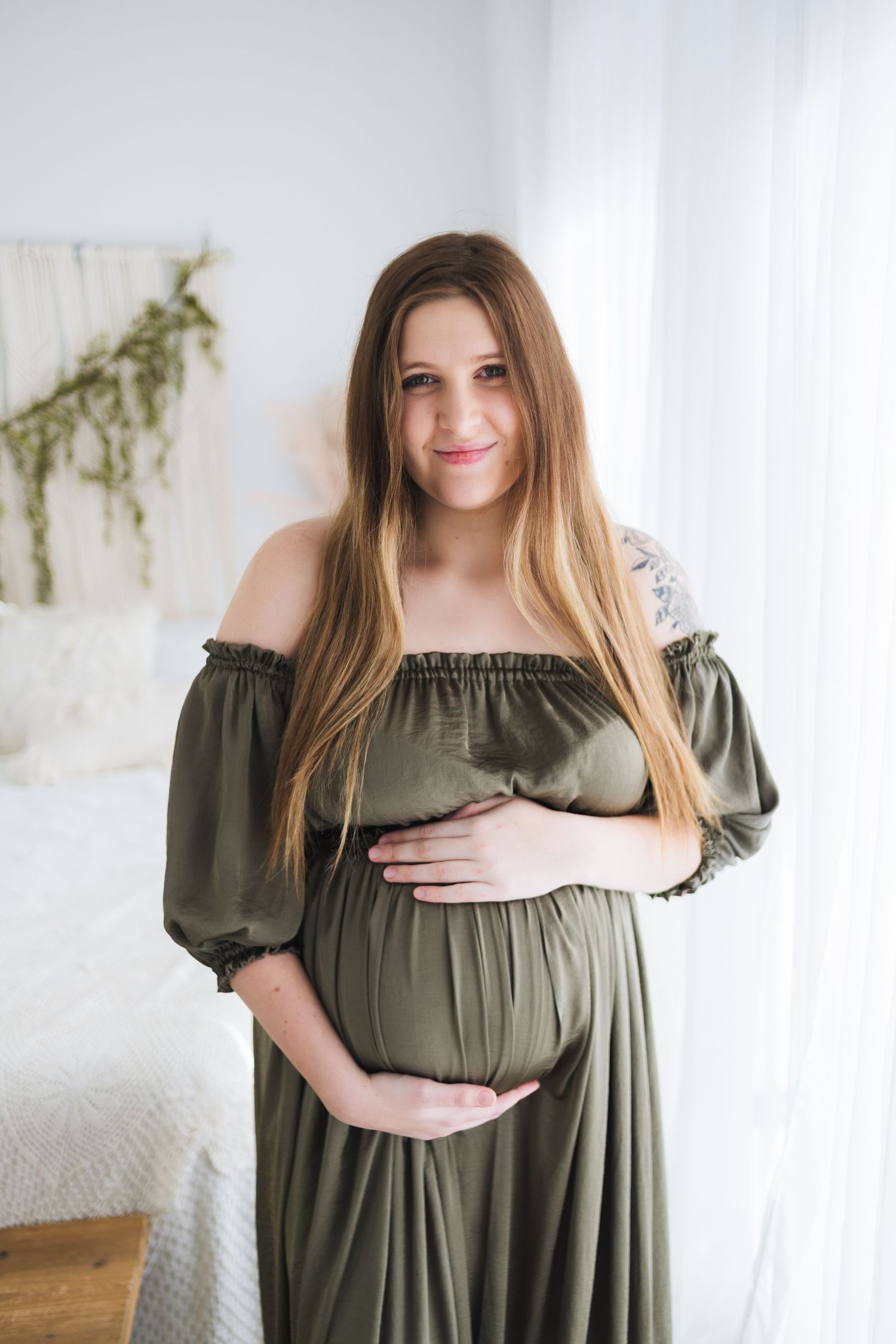Maternity portrait in olive green dress against white backdrop with natural lighting.