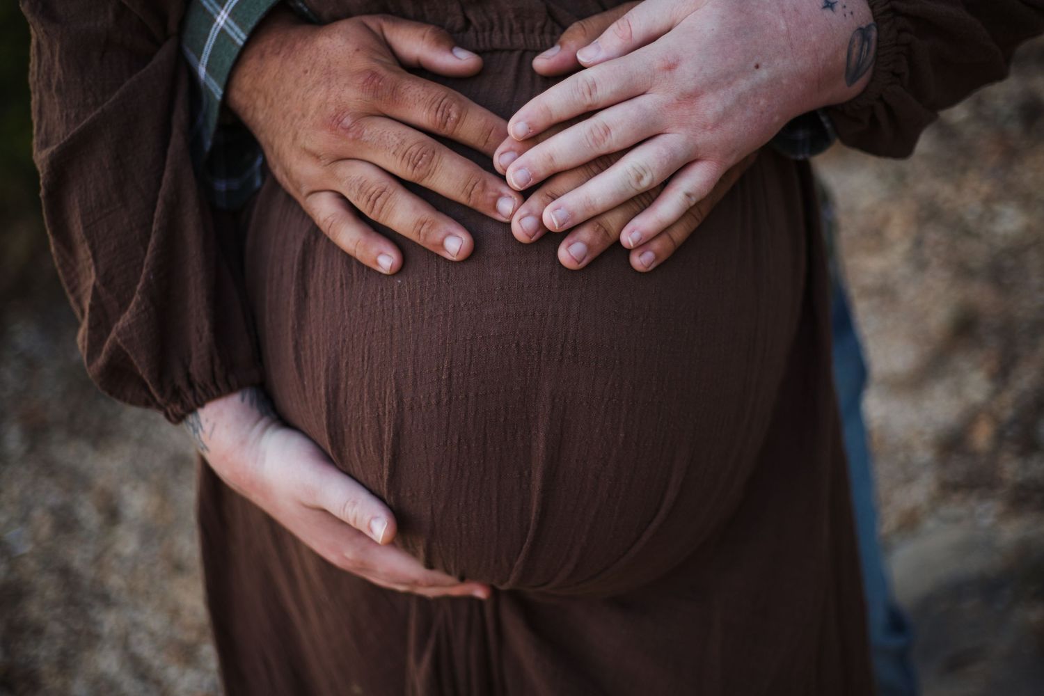 Close-up view of hands resting on a brown maternity dress showing the curves of a pregnant belly.
