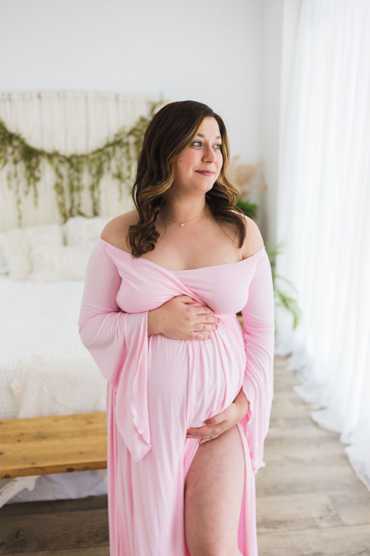 An expectant mother in a pink off-shoulder dress poses by a window with natural light.