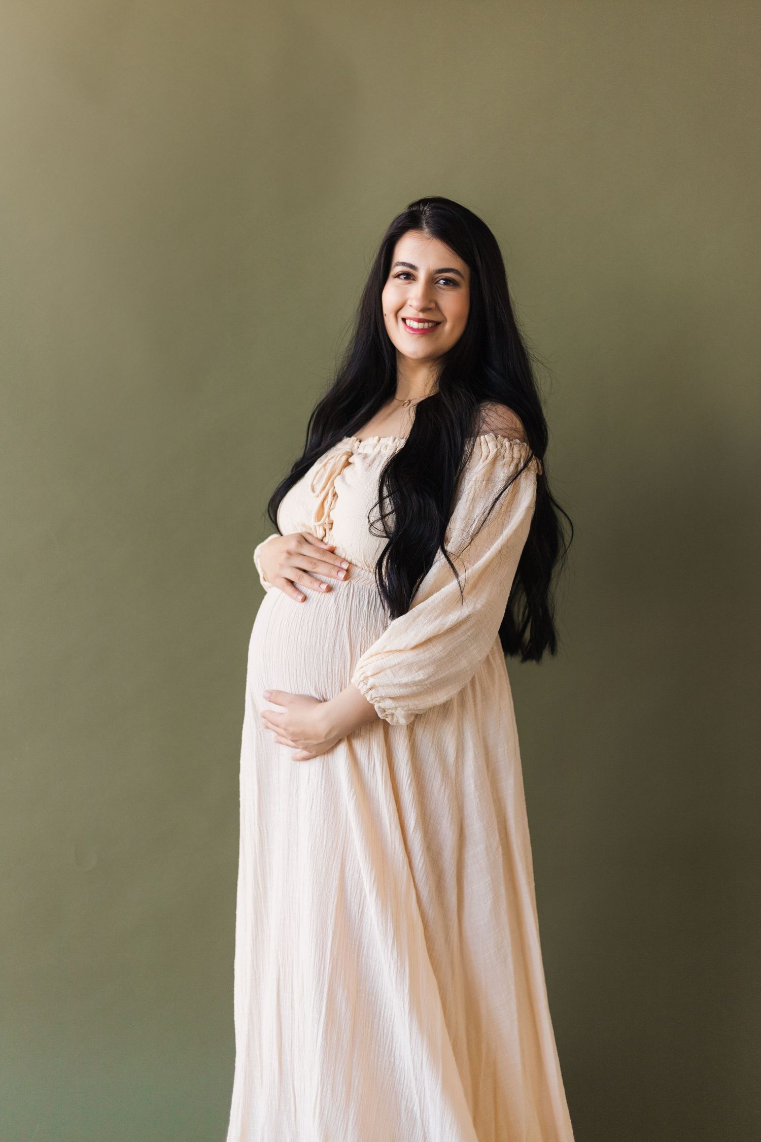 A pregnant woman in a cream-colored dress poses against an olive wall.