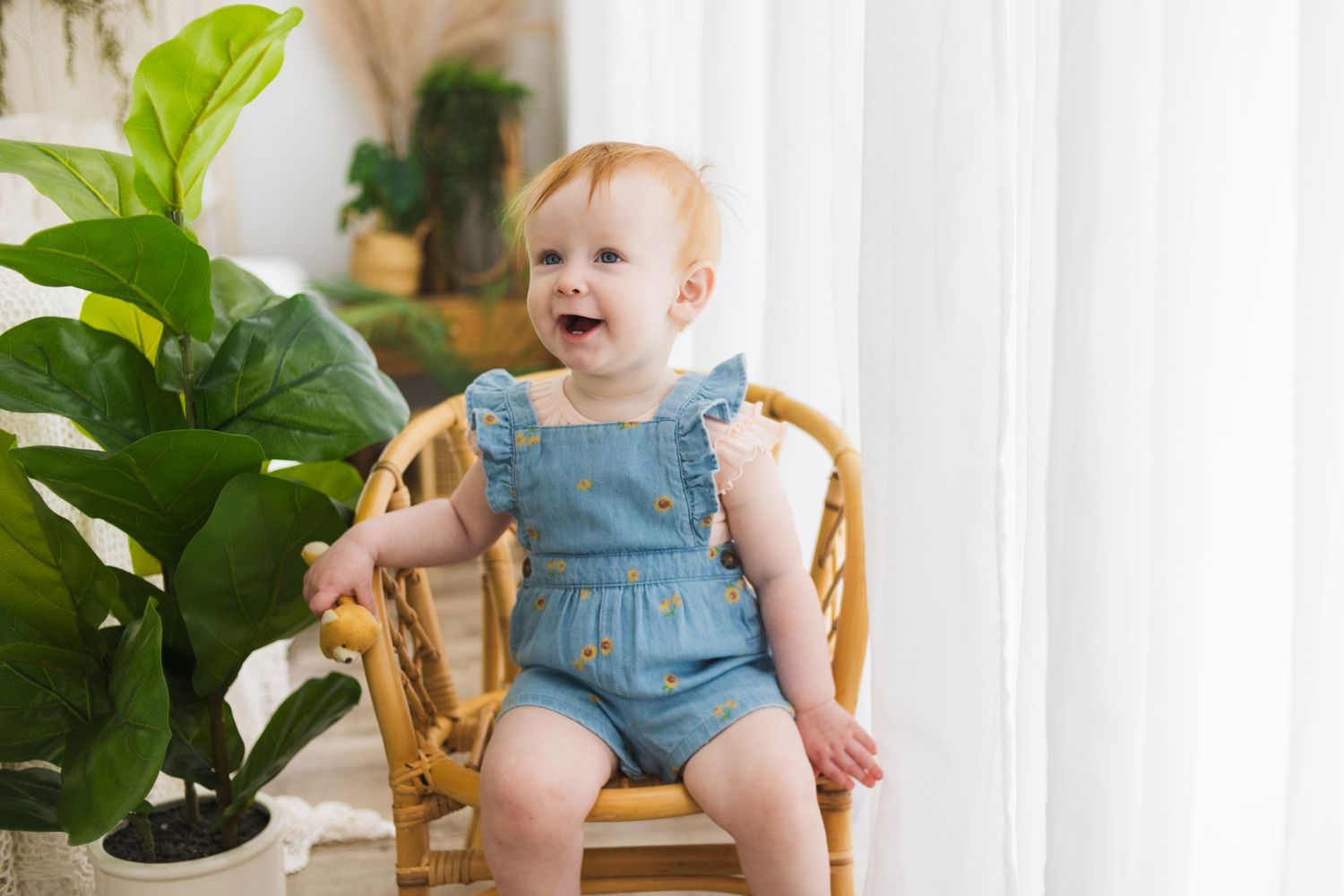 Smiling toddler in denim overalls sits in wicker chair near houseplants.