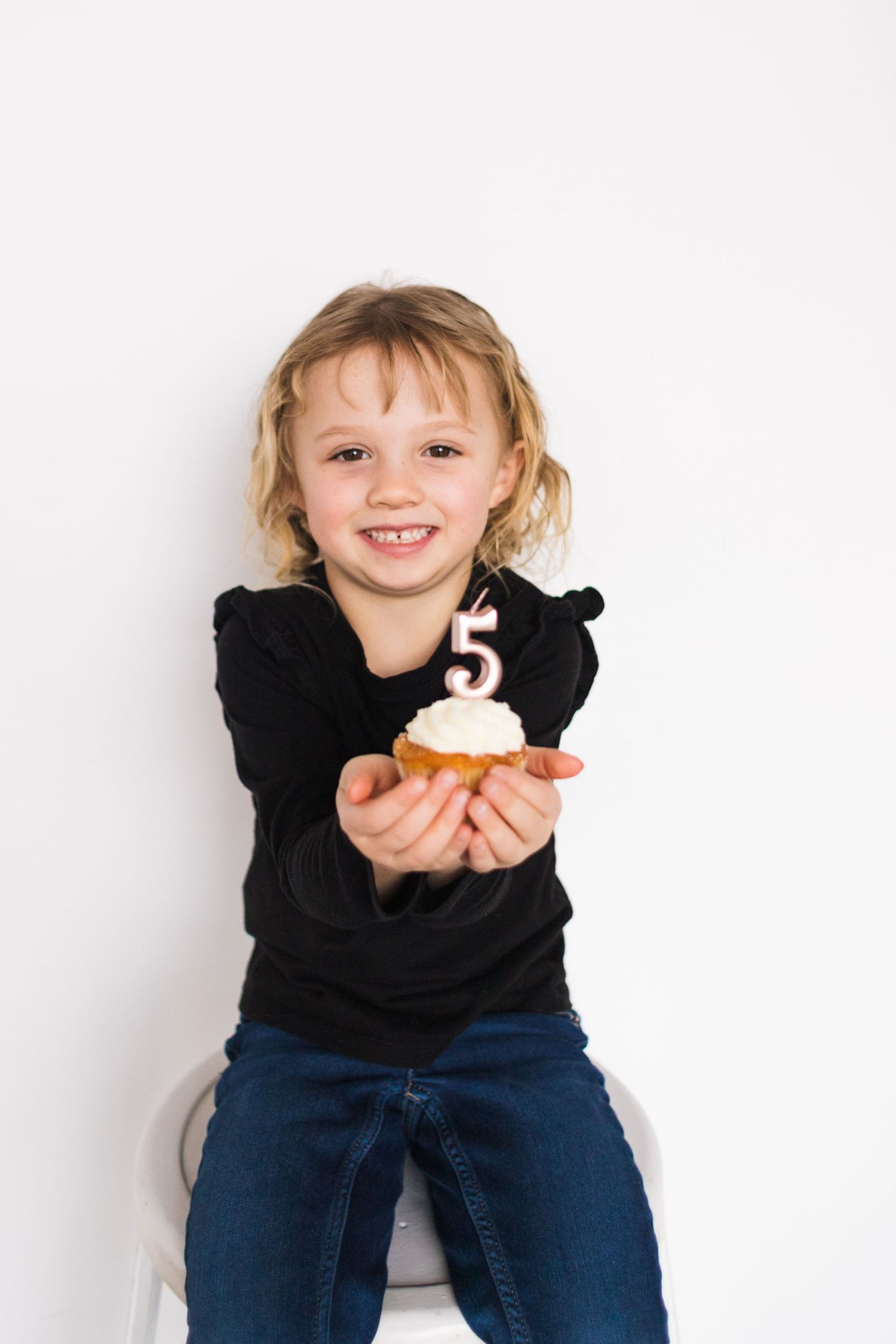 Child wearing black sweater and jeans enjoys a cupcake against white background.