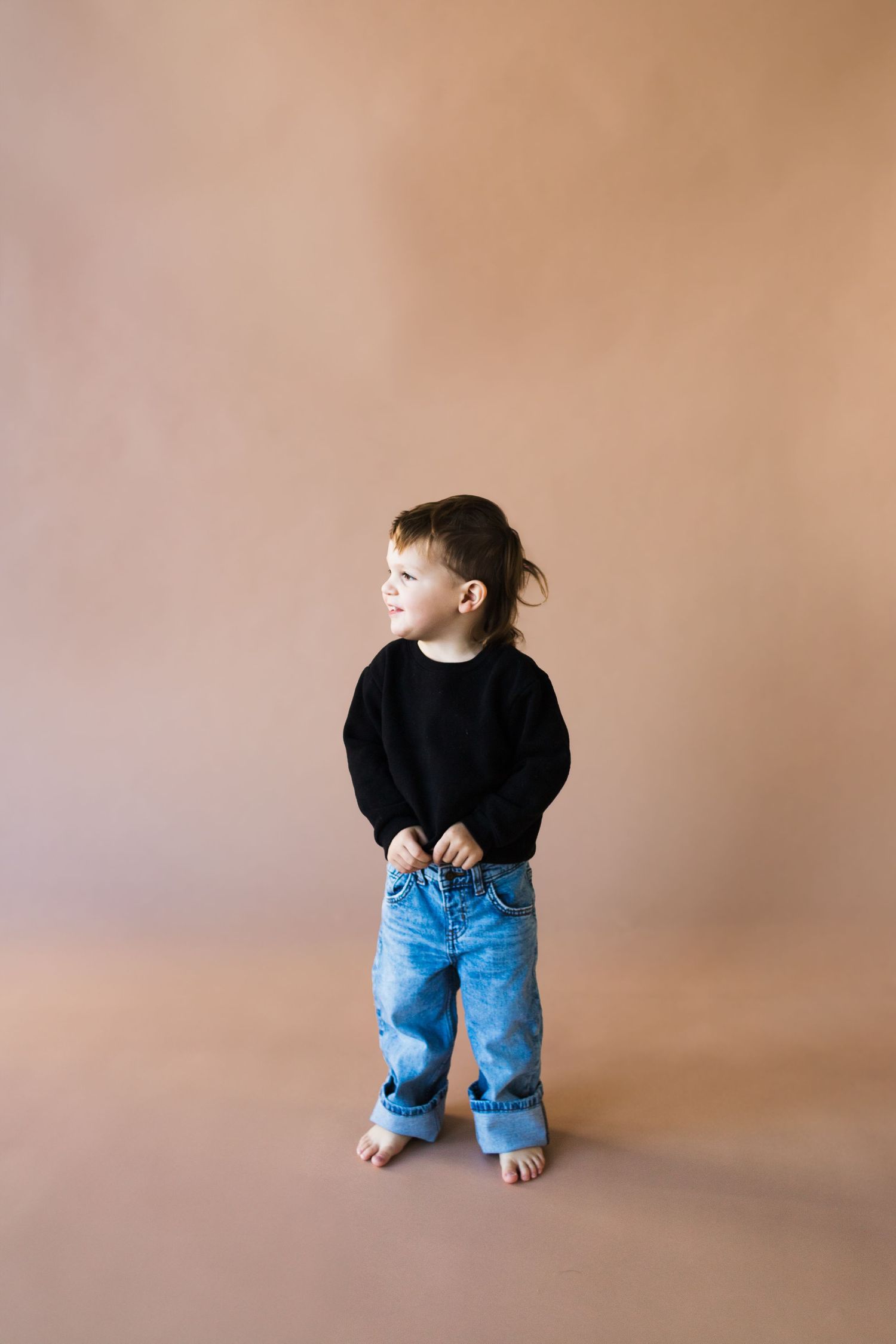 Young child in black top and jeans sits on brown studio backdrop.