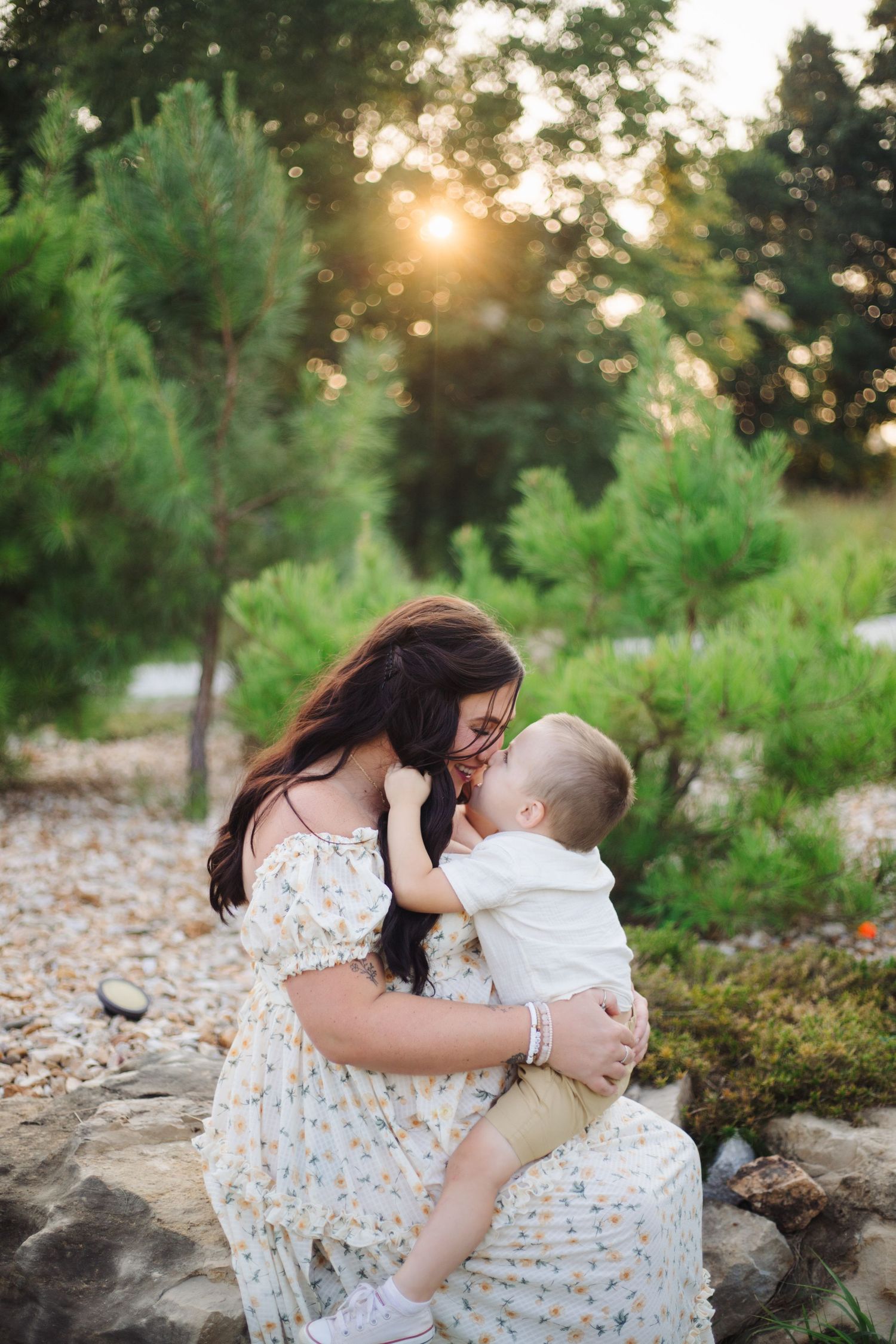 Mother and baby share tender moments at sunset in a lush outdoor setting with pine trees and rocks.