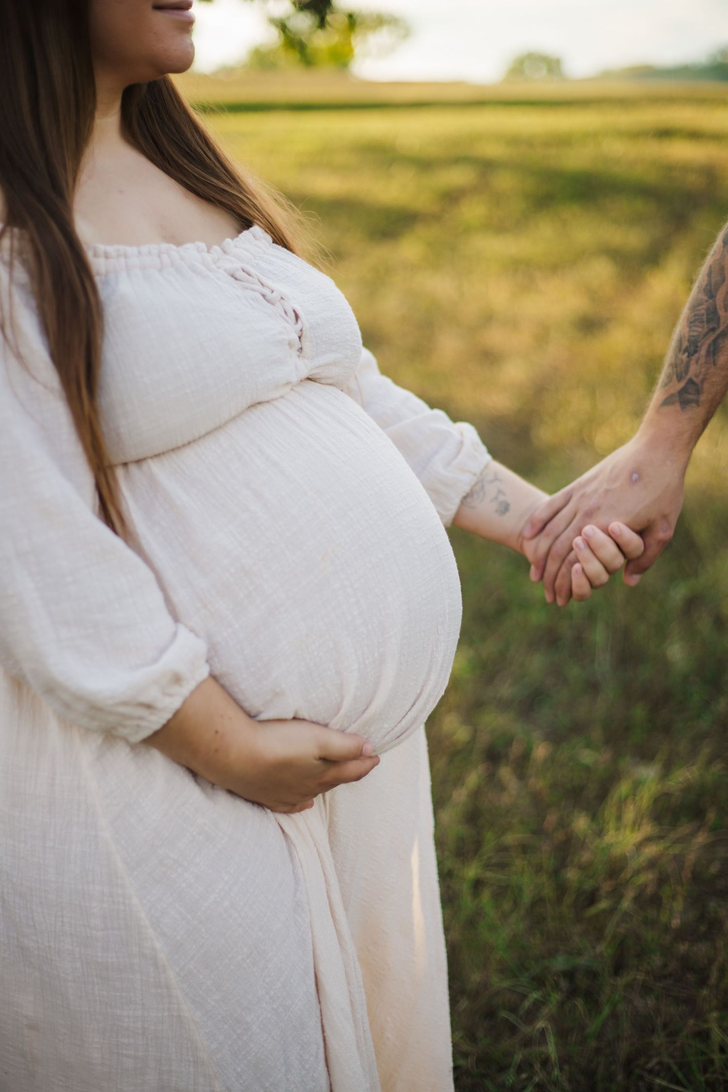 A pregnant woman in a white lace dress holds hands with someone while standing in a sunlit field at golden hour.