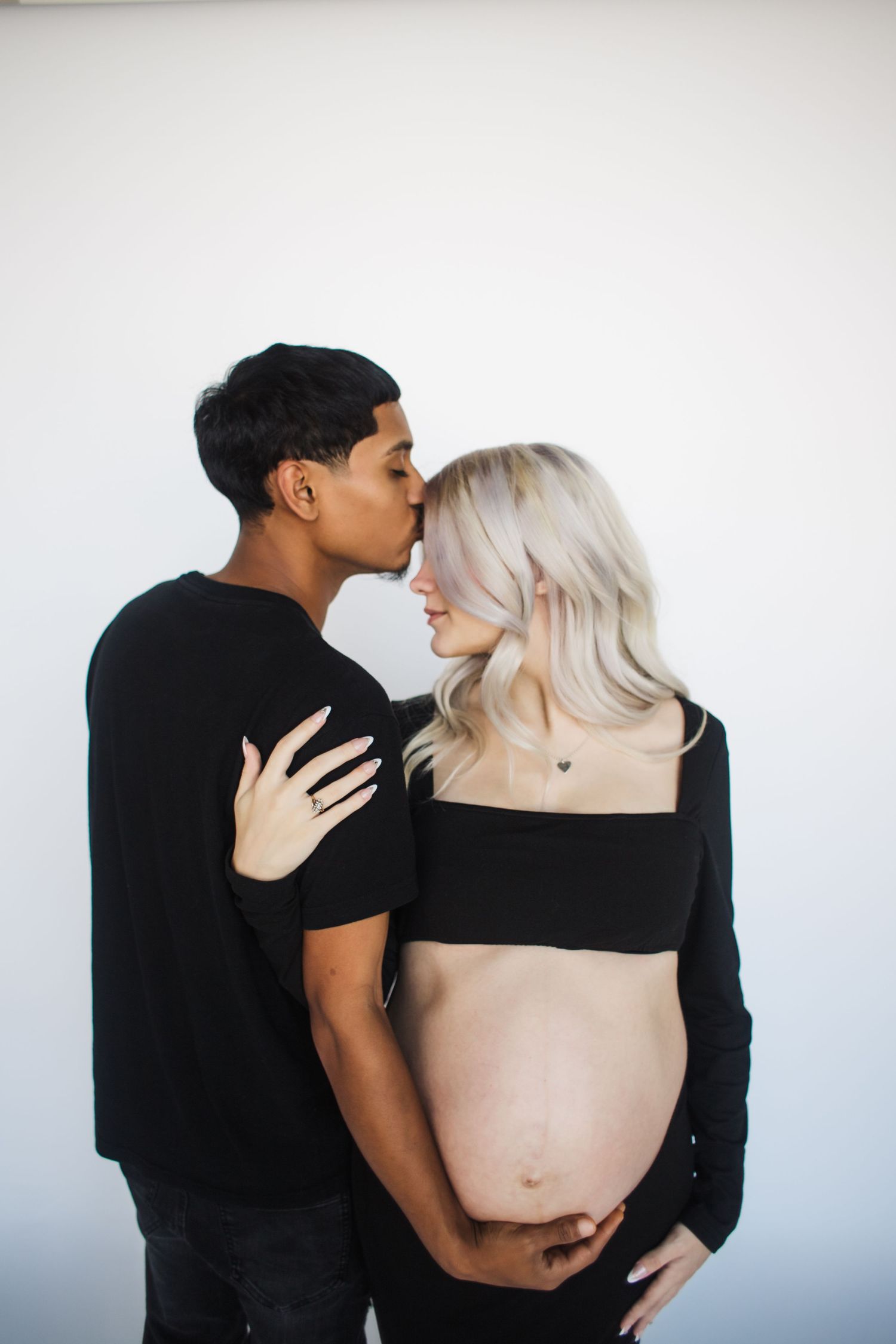 Intimate maternity photo of an expectant couple in black outfits against a white wall.