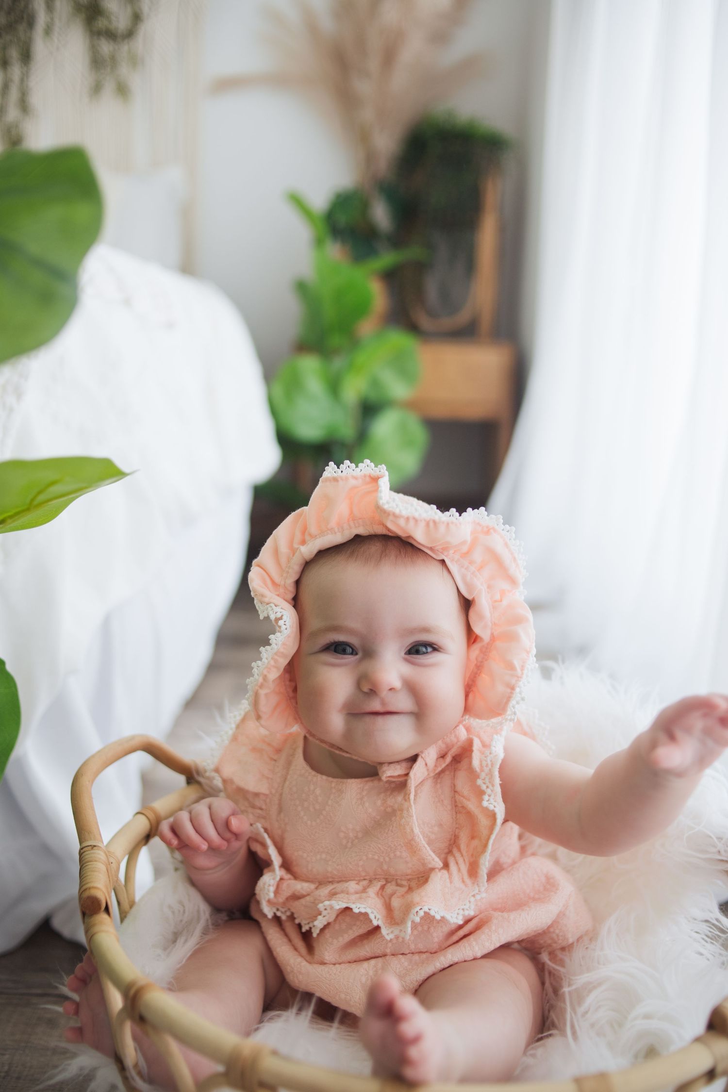Baby wearing a delicate peach-colored ruffled outfit sits on white fabric with greenery in the background.
