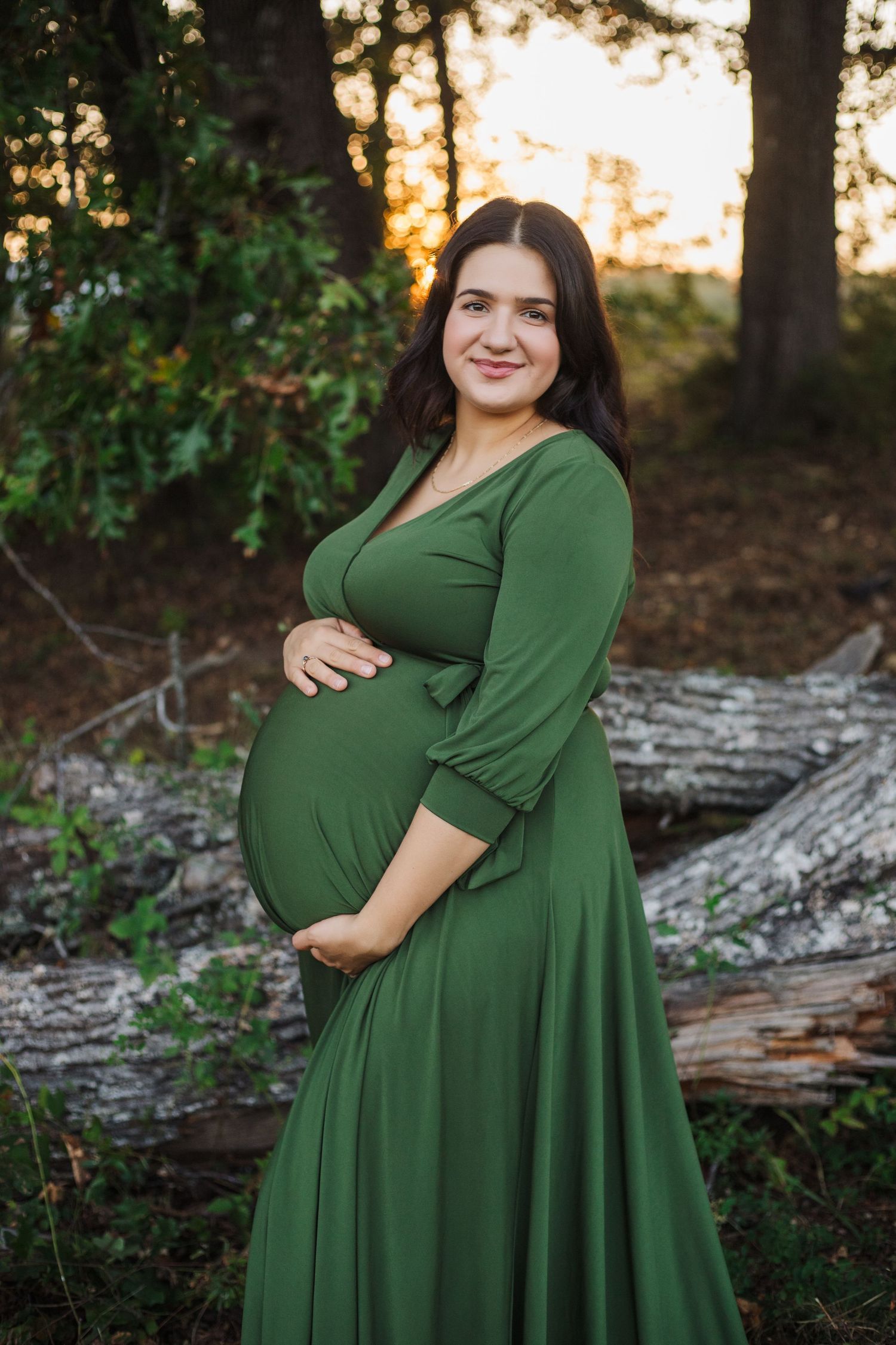 A woman in a forest green dress poses in a wooded setting at sunset during a maternity photoshoot.