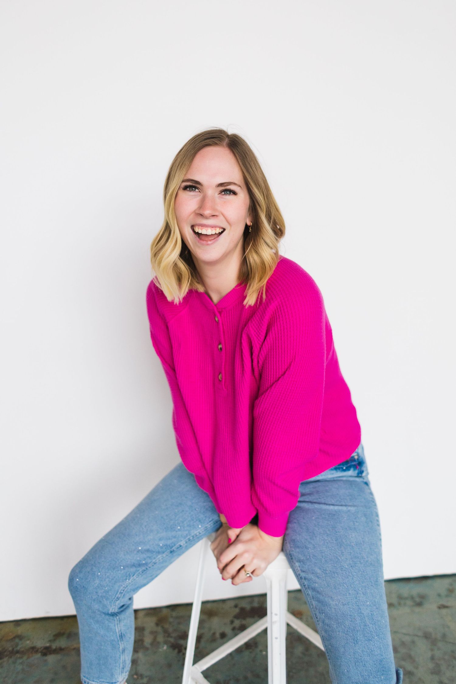 Person in vibrant magenta sweater poses cheerfully on stool against white wall.