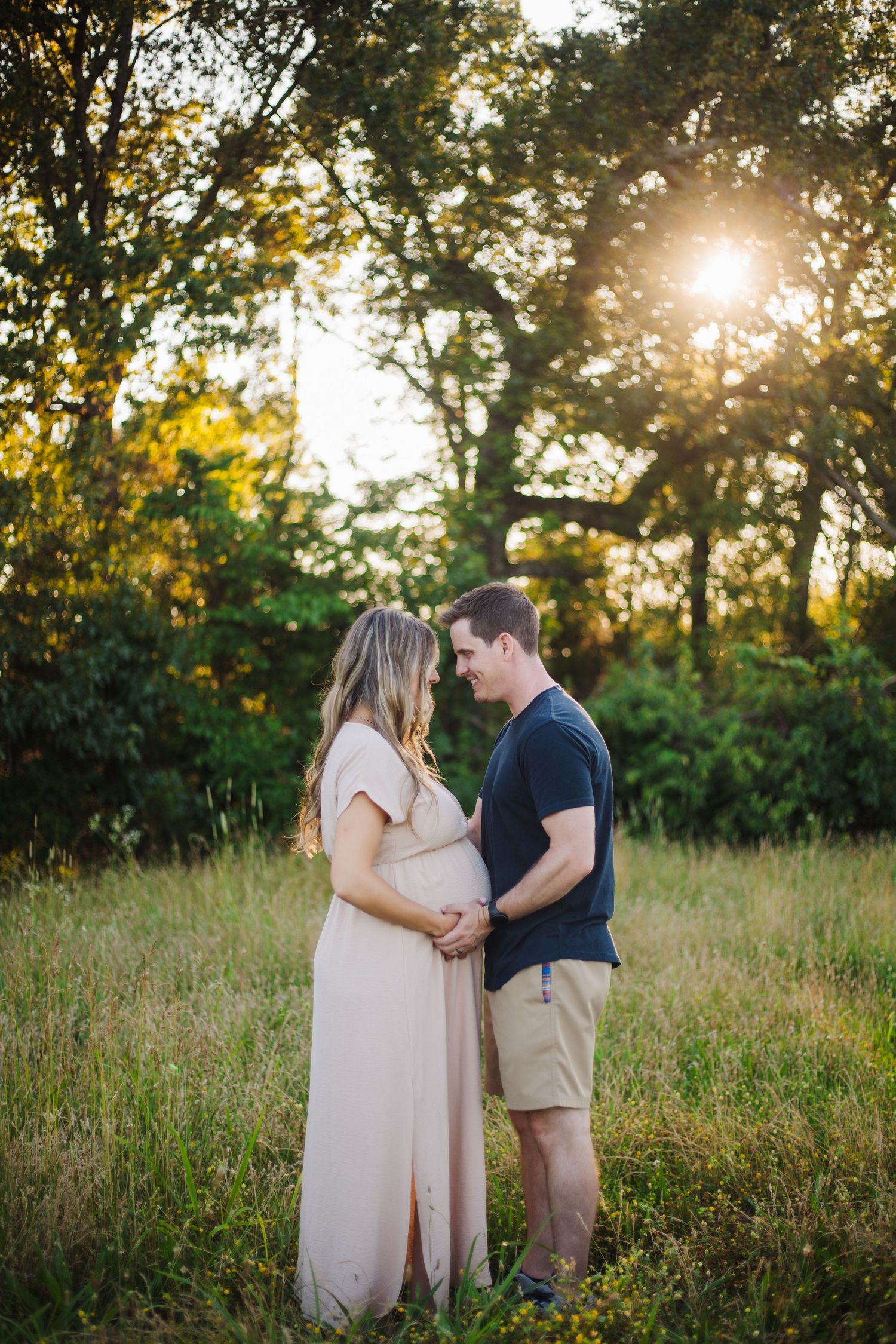 A couple shares an intimate moment in a sunlit meadow during a maternity photoshoot at golden hour.