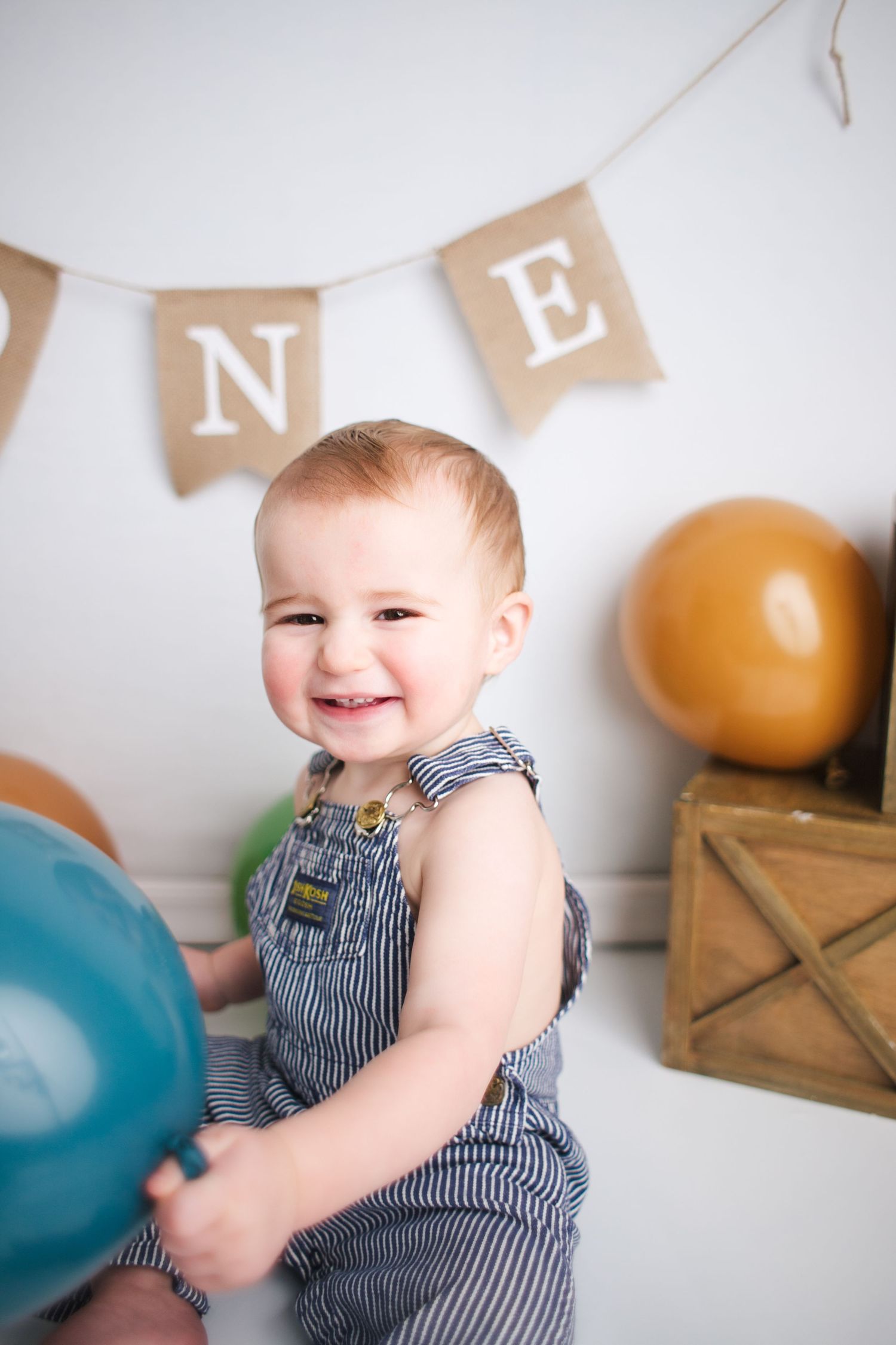 A happy baby in striped overalls sits and plays with blue and beige balloons during a photo session.