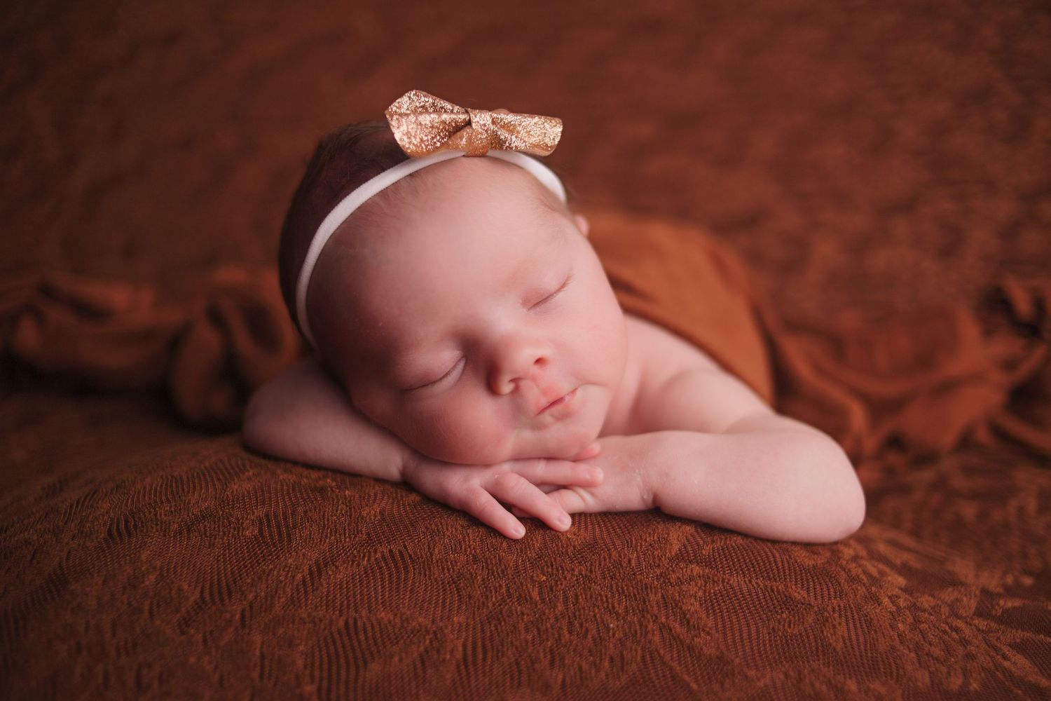 Sleeping newborn baby wearing a peach bow headband rests peacefully on brown textured fabric.
