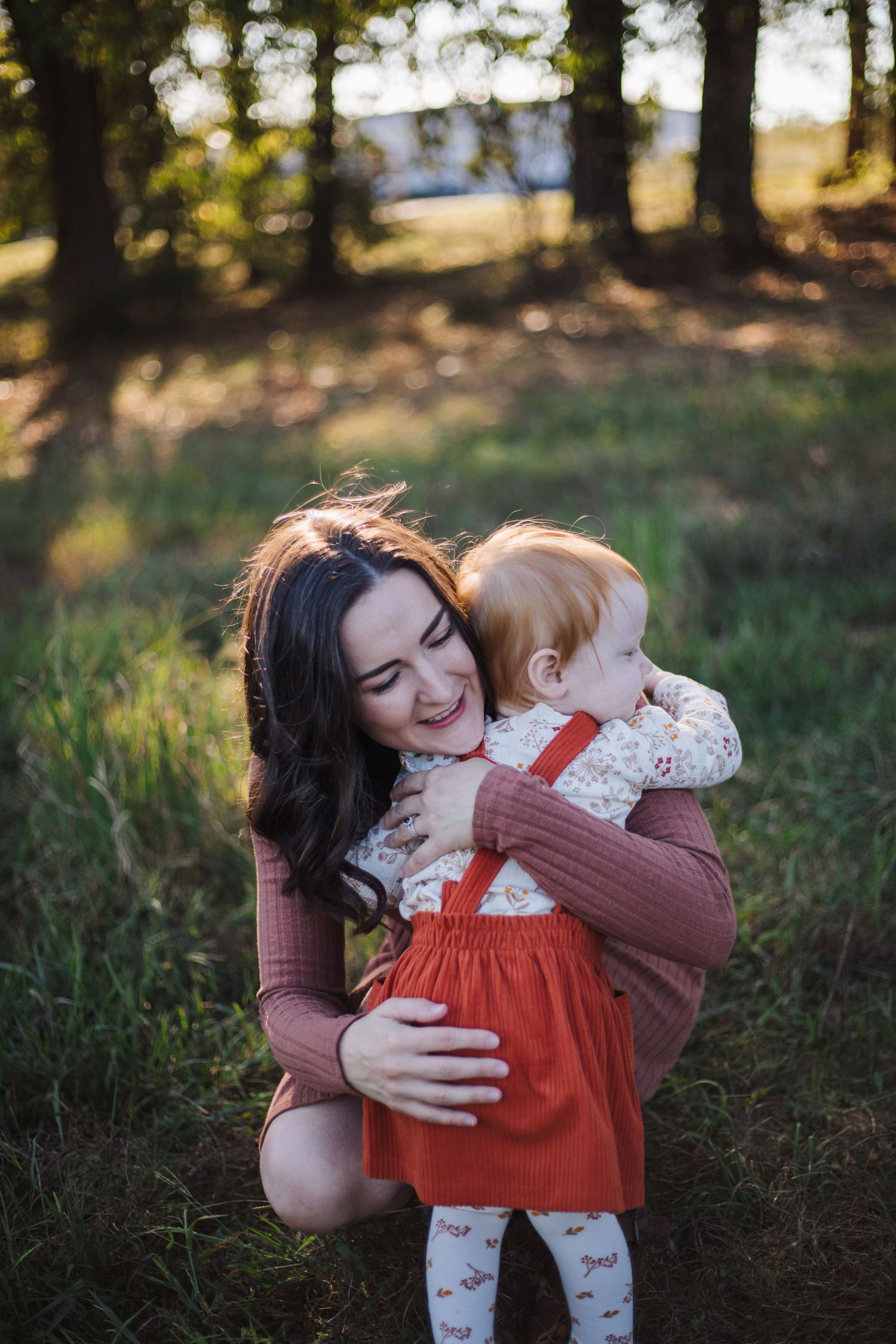 A mother in a brown sweater lovingly embraces her young child wearing an orange dress in a sunlit forest setting.