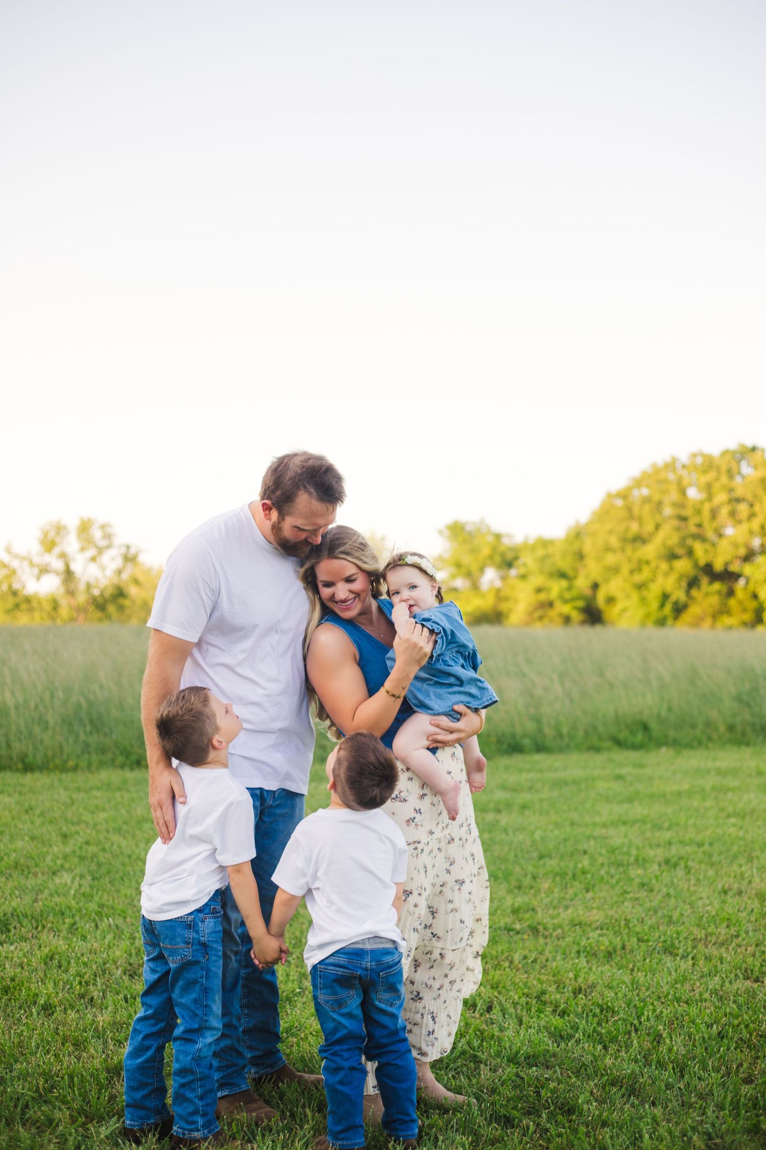 A happy family of five poses outdoors in a green field, with parents embracing three young children at golden hour.