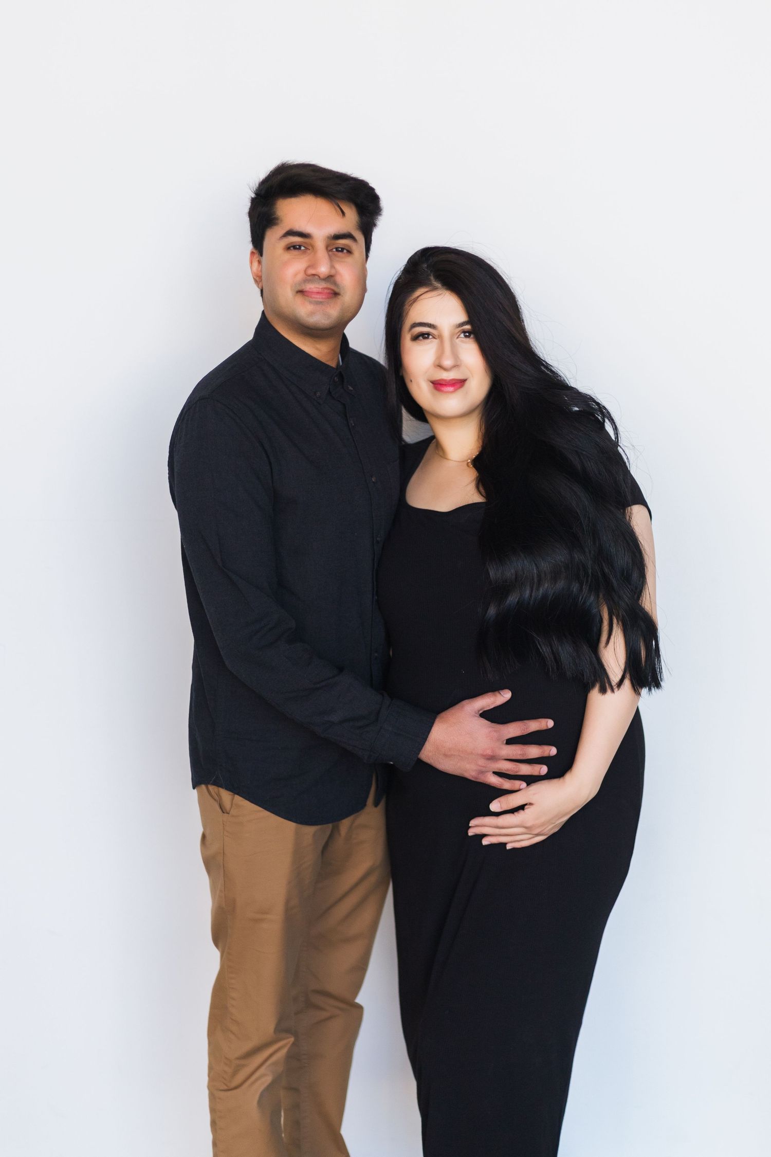 Couple in black outfits embrace while posing against white studio background.