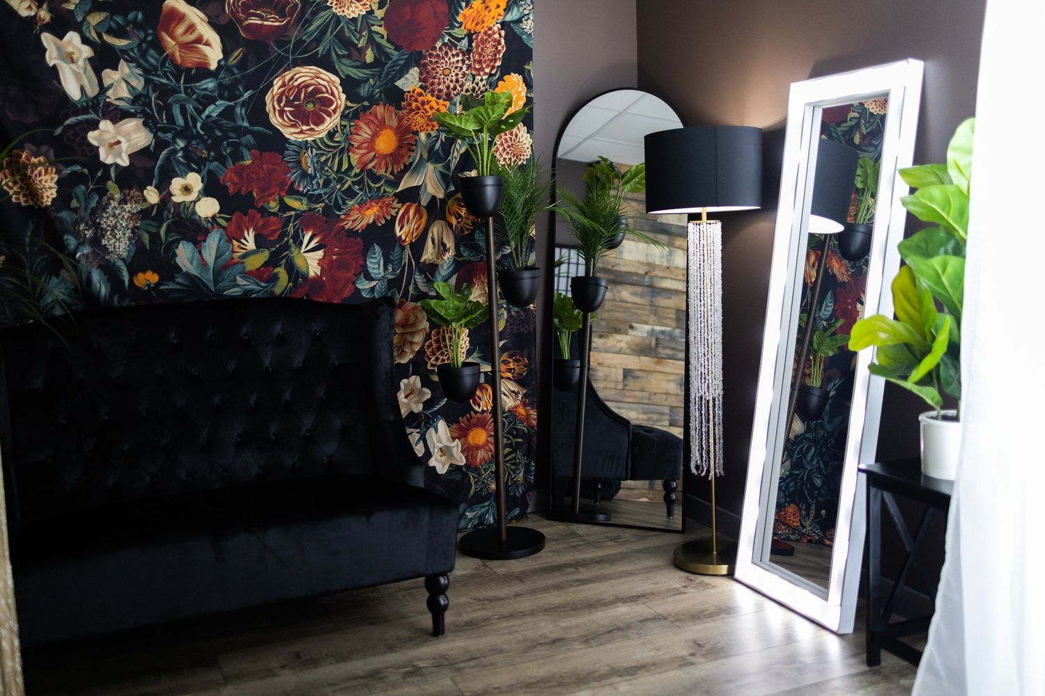 Modern photo studio with dark floral wallpaper, black velvet sofa, and full-length mirror.