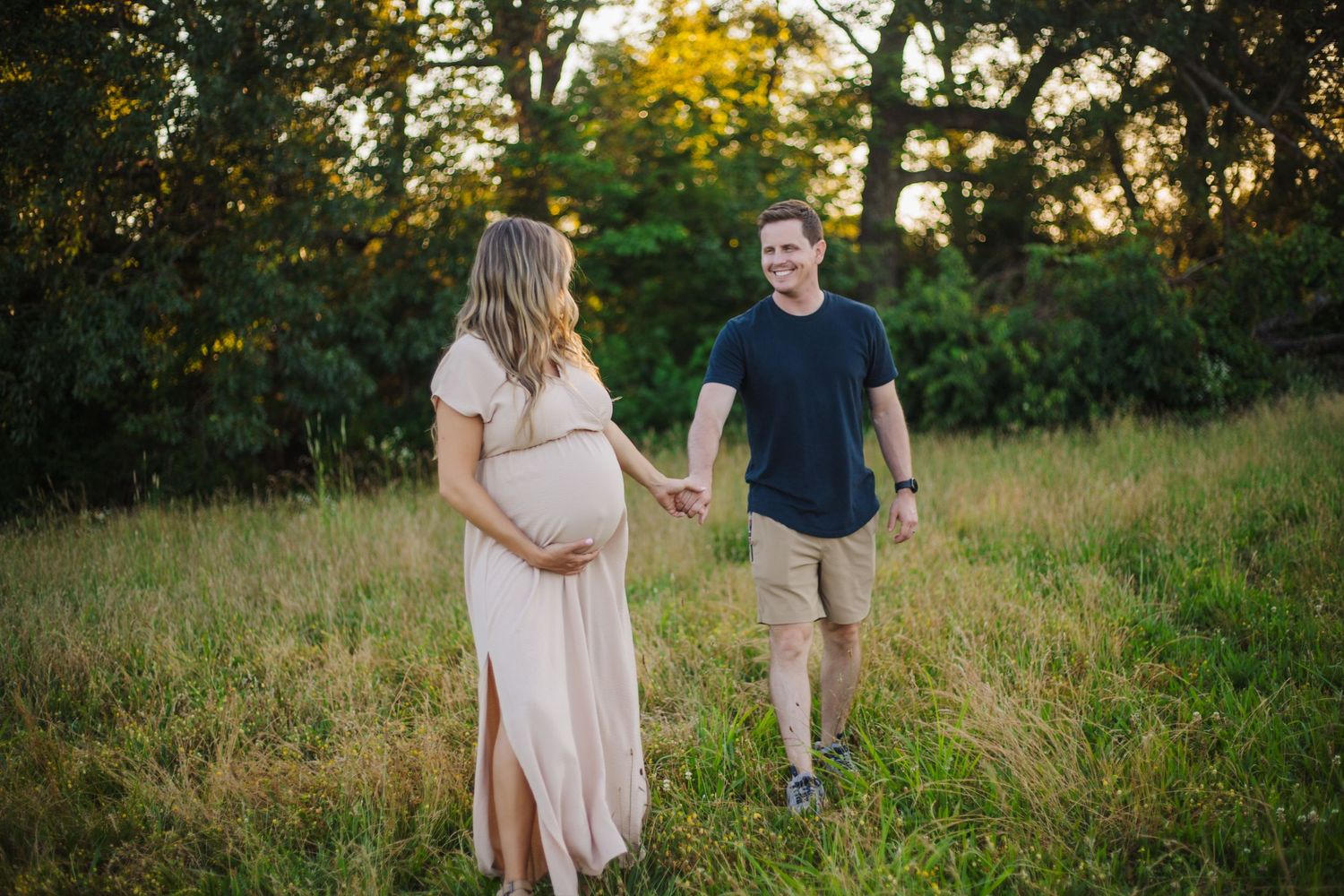 Maternity photo of a couple walking through a sunlit field at golden hour.
