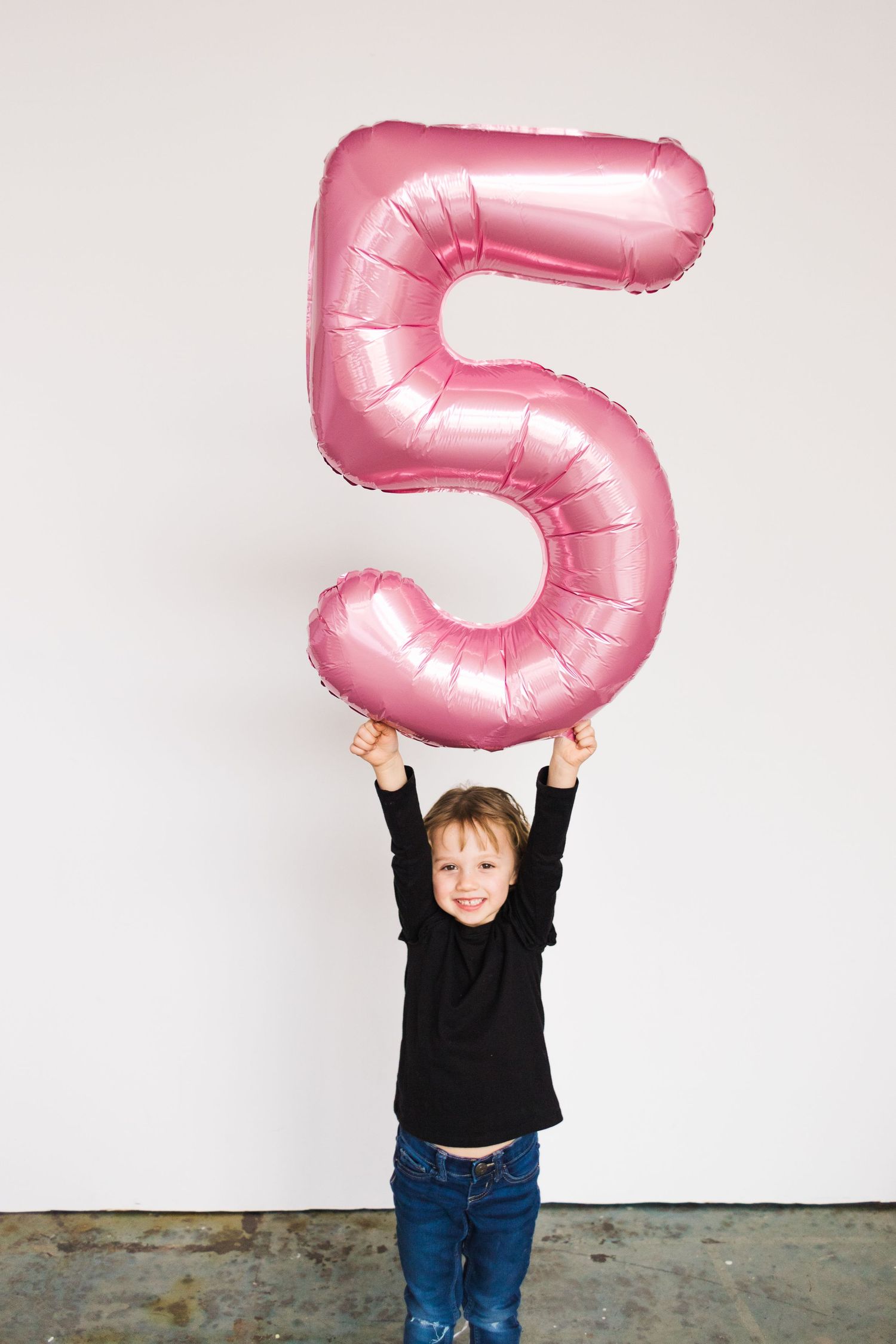 Child holds large pink number 5 balloon against white wall.