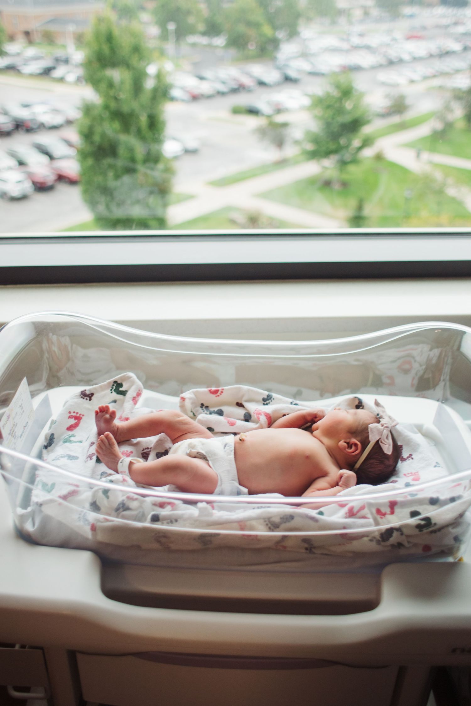 A newborn baby rests in a hospital bassinet by a window overlooking a parking lot on a bright day.