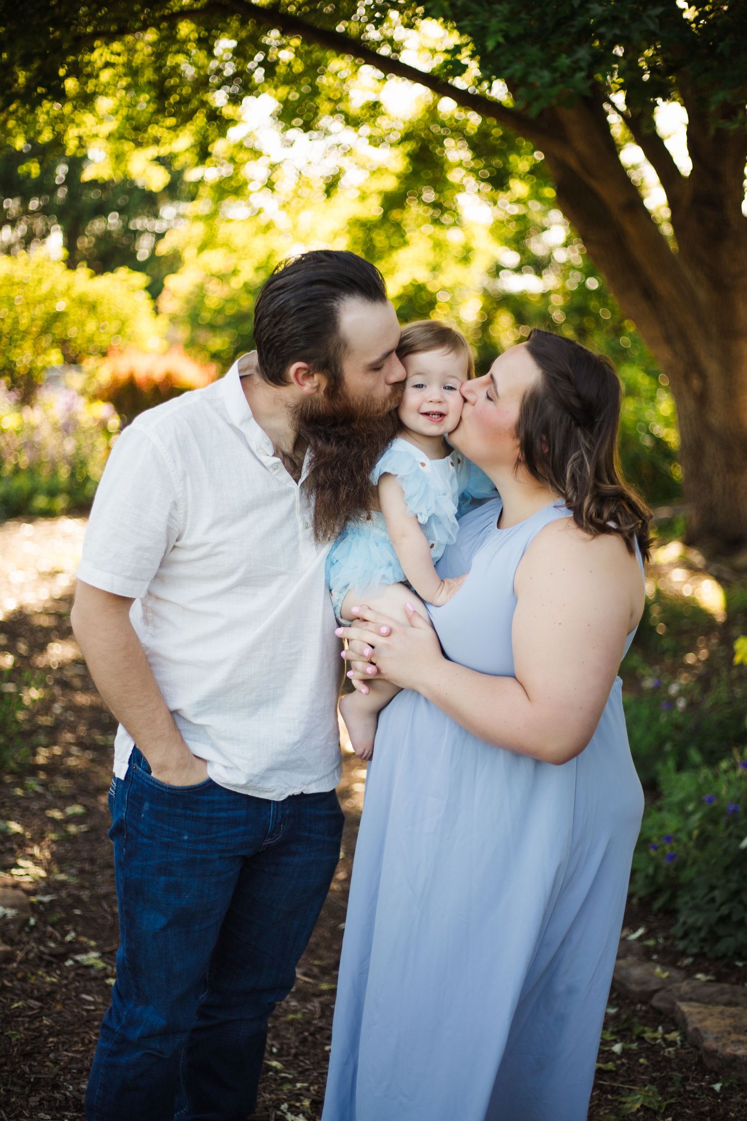 Bearded father and pregnant mother kissing toddler girl in blue dress outdoors in lush green garden setting.