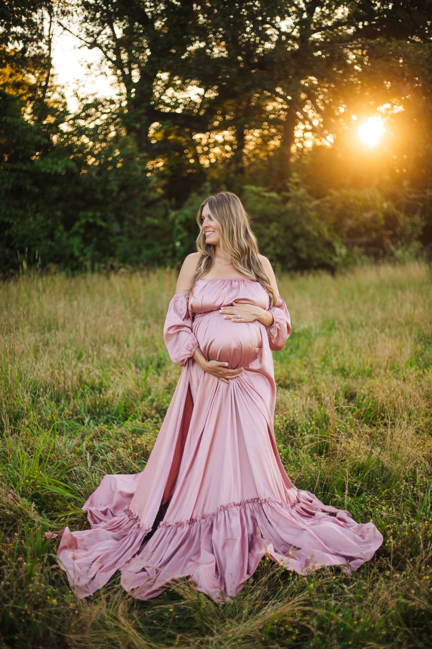 Flowing pink maternity gown photographed in golden sunset light in grassy meadow.