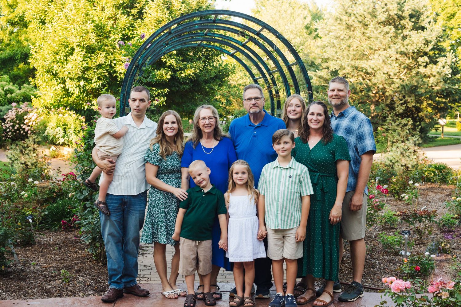 A group of people pose together under a metal garden arch surrounded by lush greenery and flowers in a park.