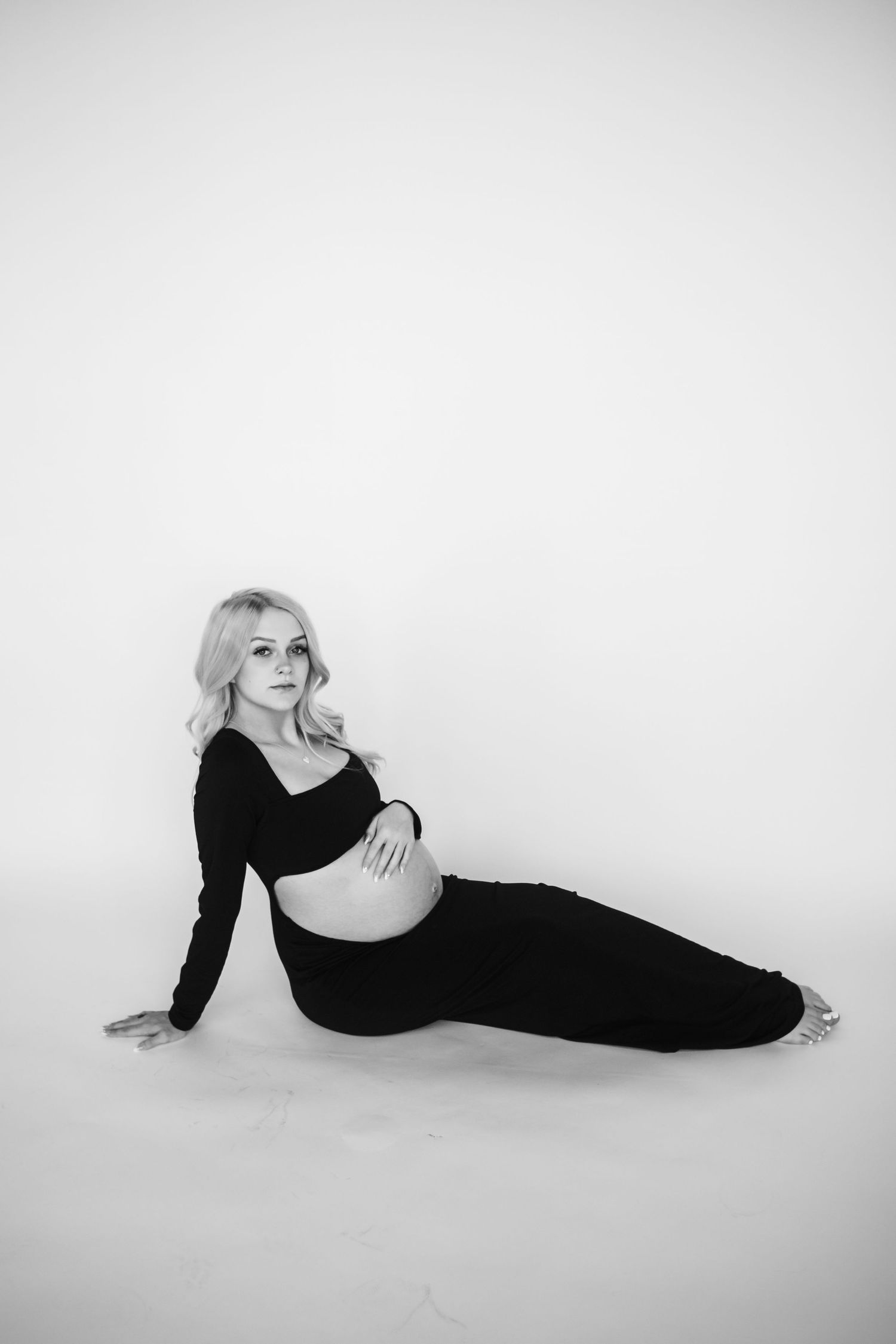 Black and white maternity photo showing a pregnant woman in dark clothing reclining against a light background.