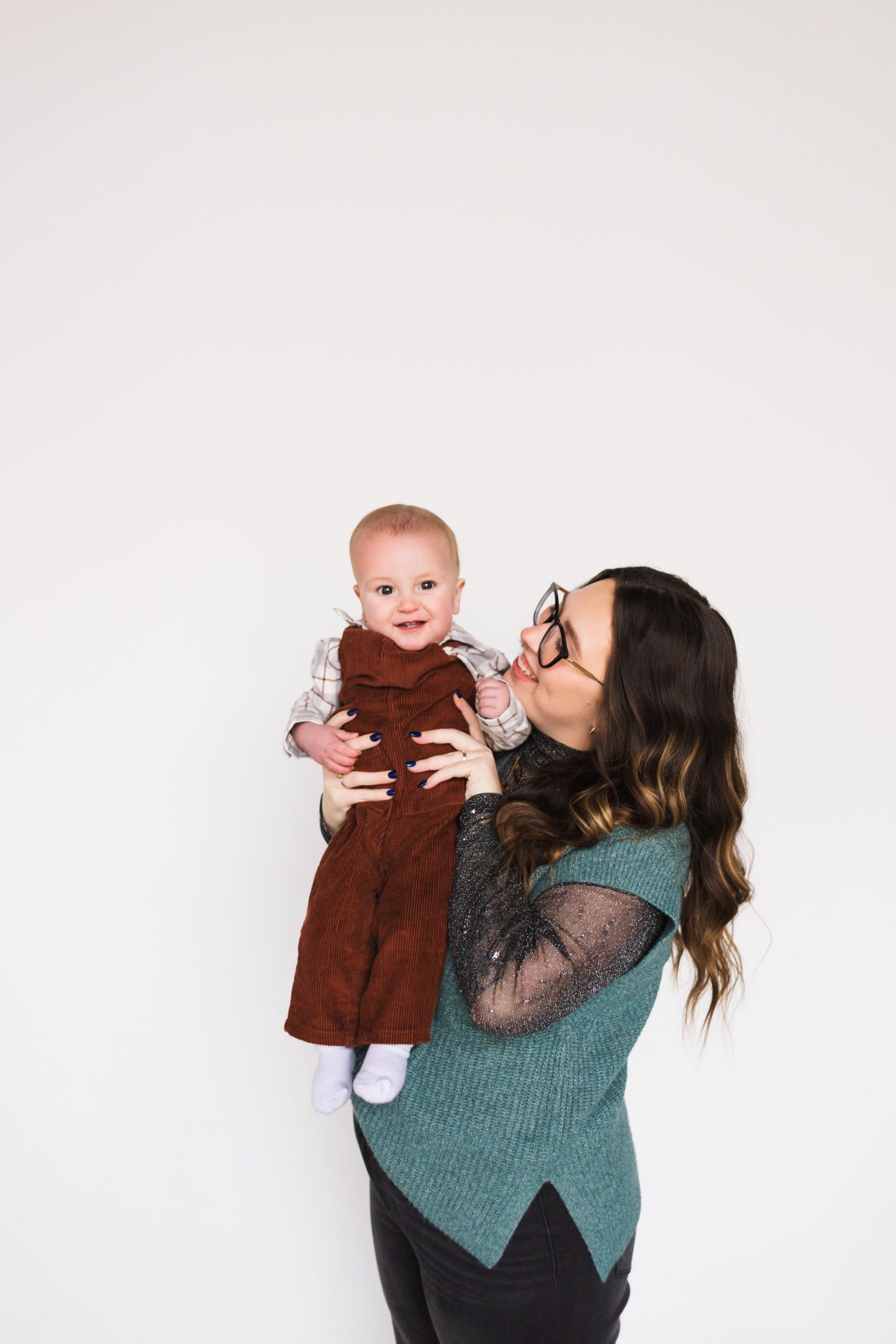 Parent in teal sweater lifts and plays with baby wearing brown overalls.