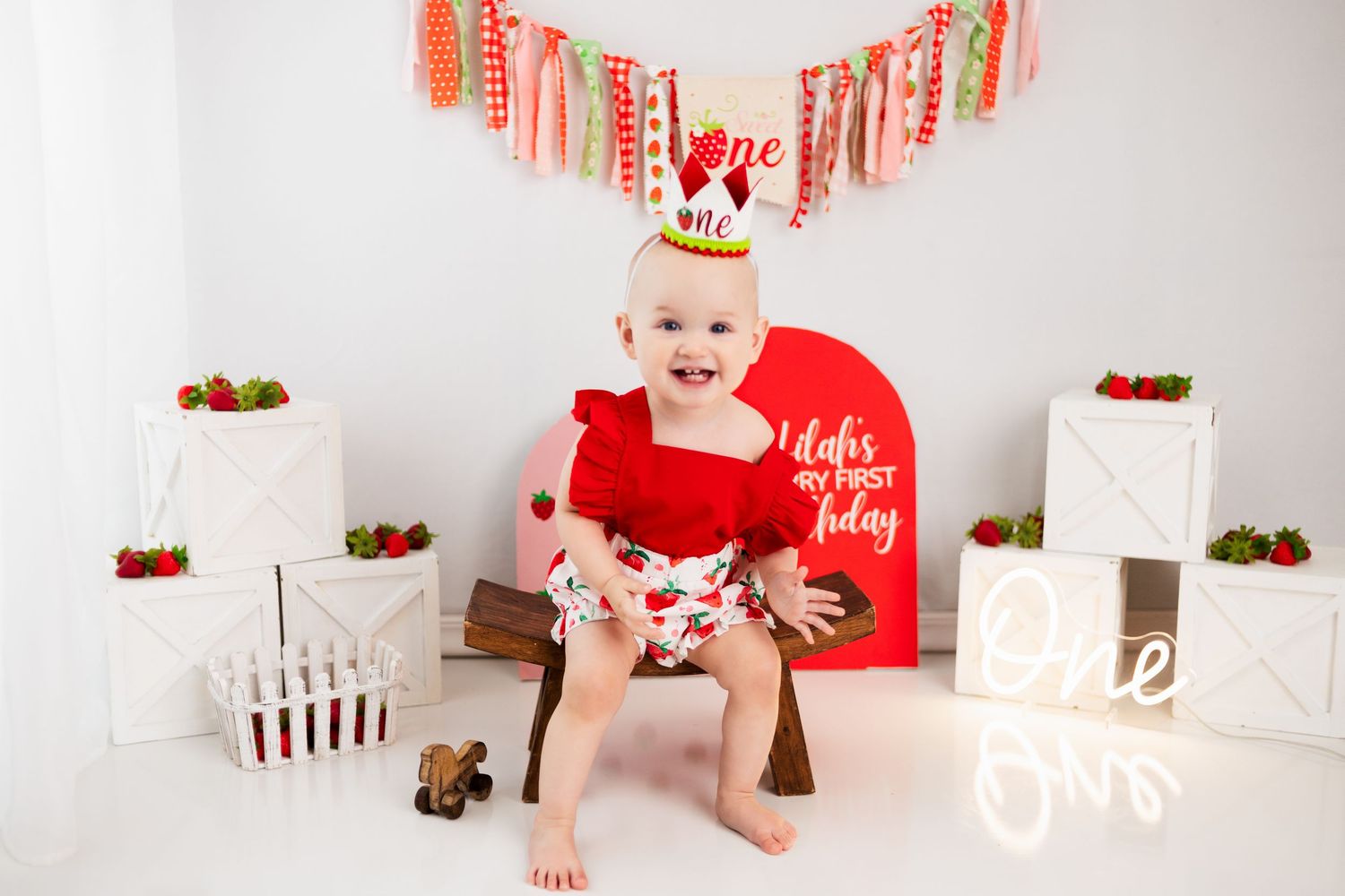 Festive first birthday setup with red decorations, strawberries, and white pedestals against light background.