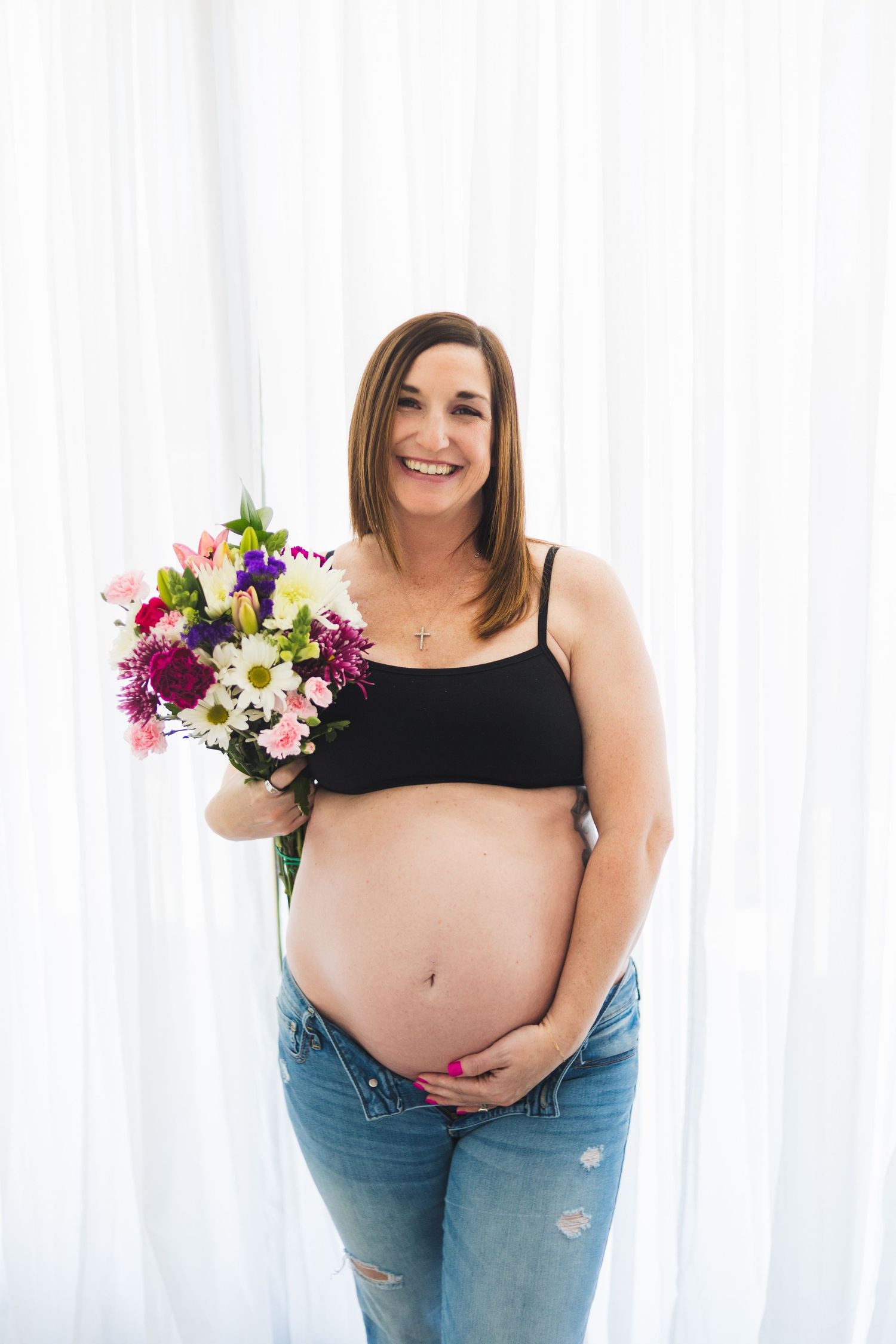 An expectant mother holds a vibrant bouquet of purple and white flowers while posing in casual attire.