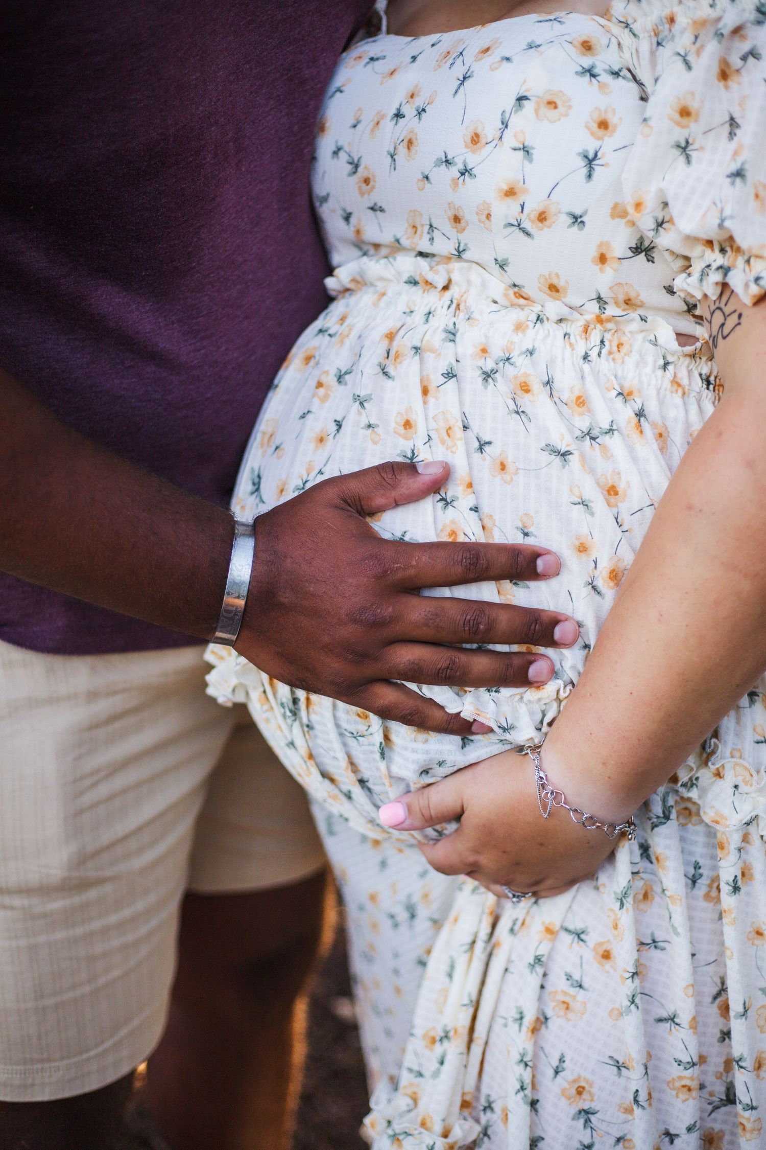 Close-up of hands tenderly touching a pregnant belly wearing a white floral dress against a purple background.