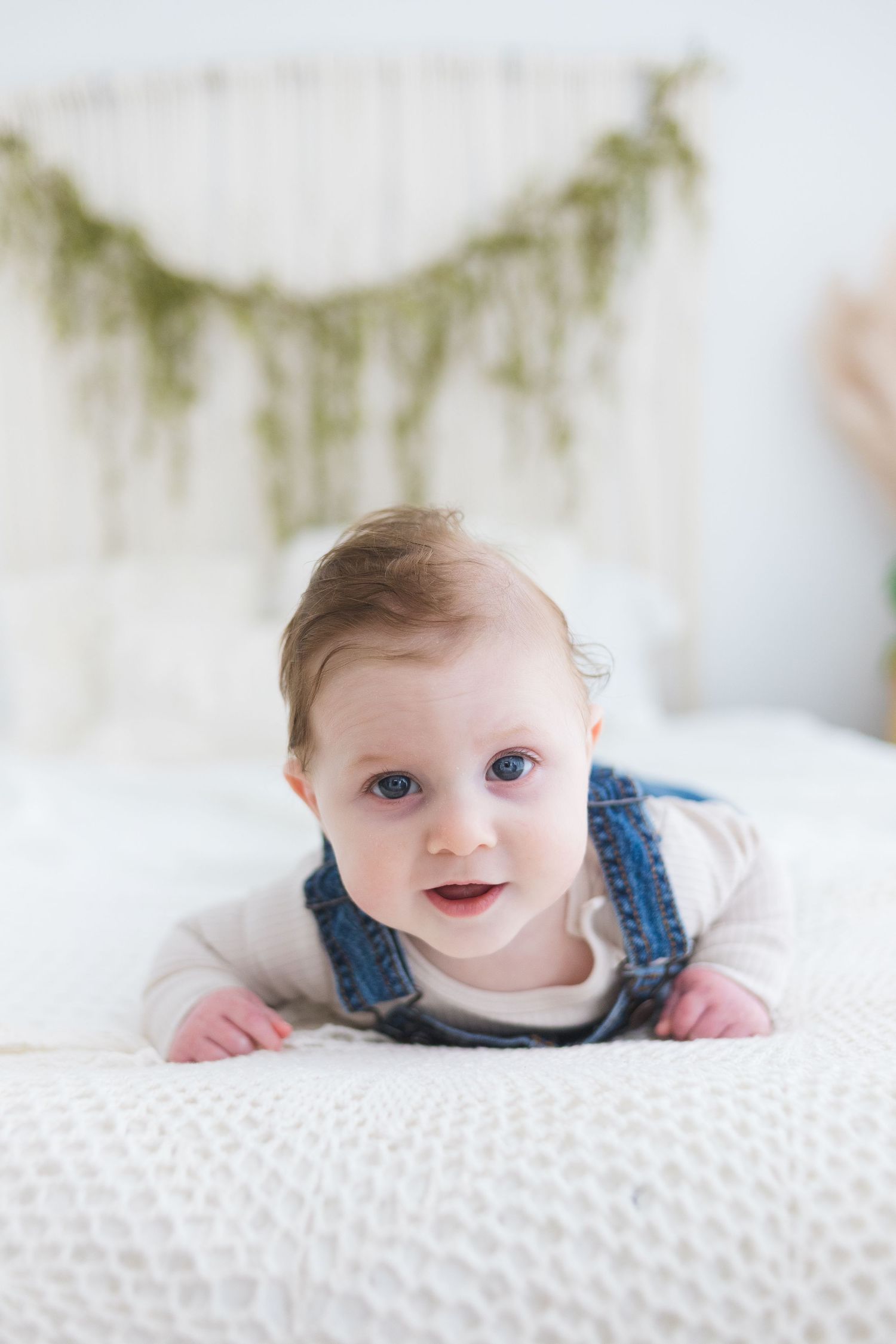 A baby in denim overalls lies on a white textured surface smiling at the camera in a series of three photos.